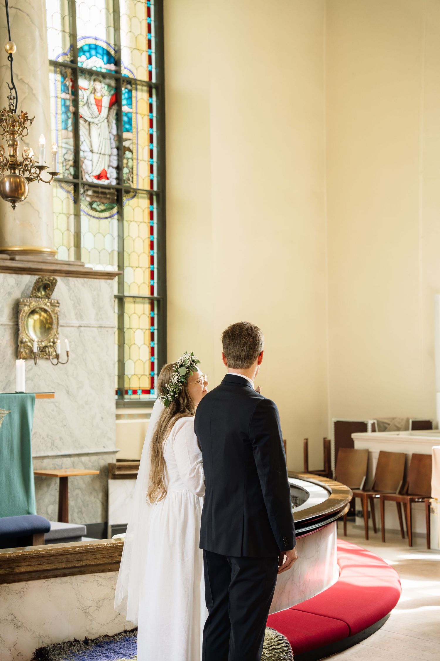 Bride and groom stand together at church altar admiring stained glass window during wedding.