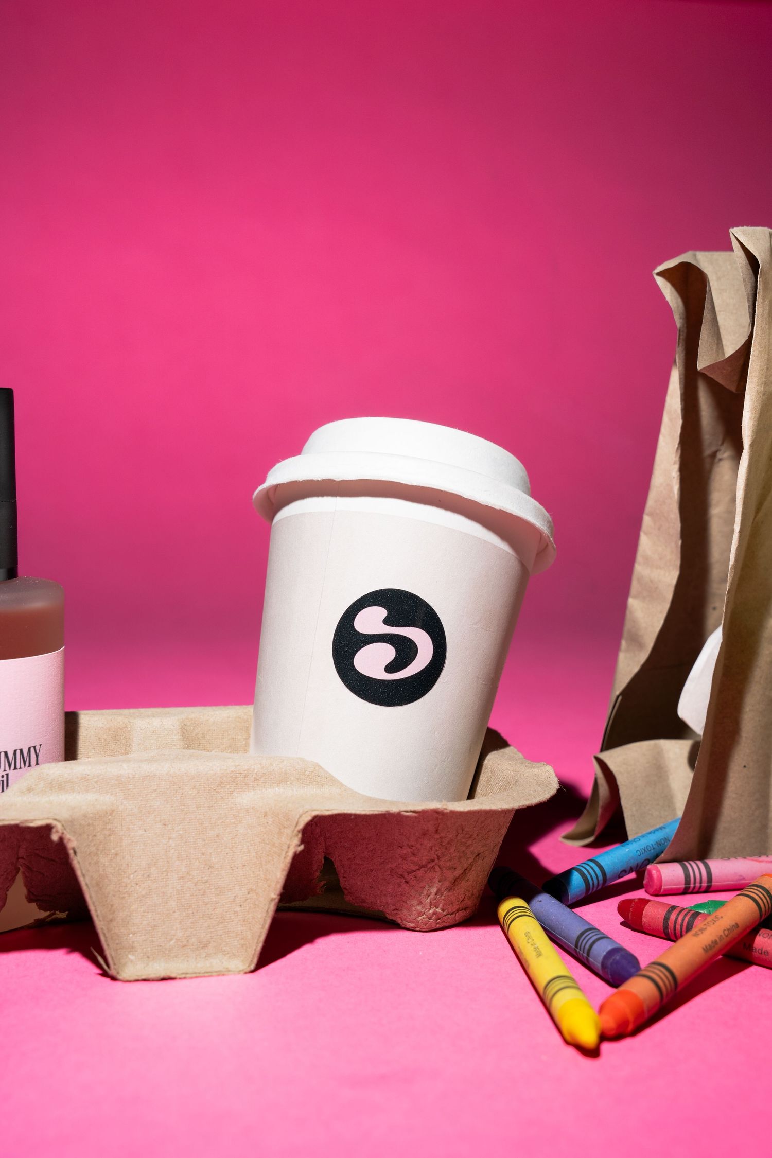 White coffee cup with logo in cardboard holder, crayons, paper bag, and pink bottle on vibrant pink background.