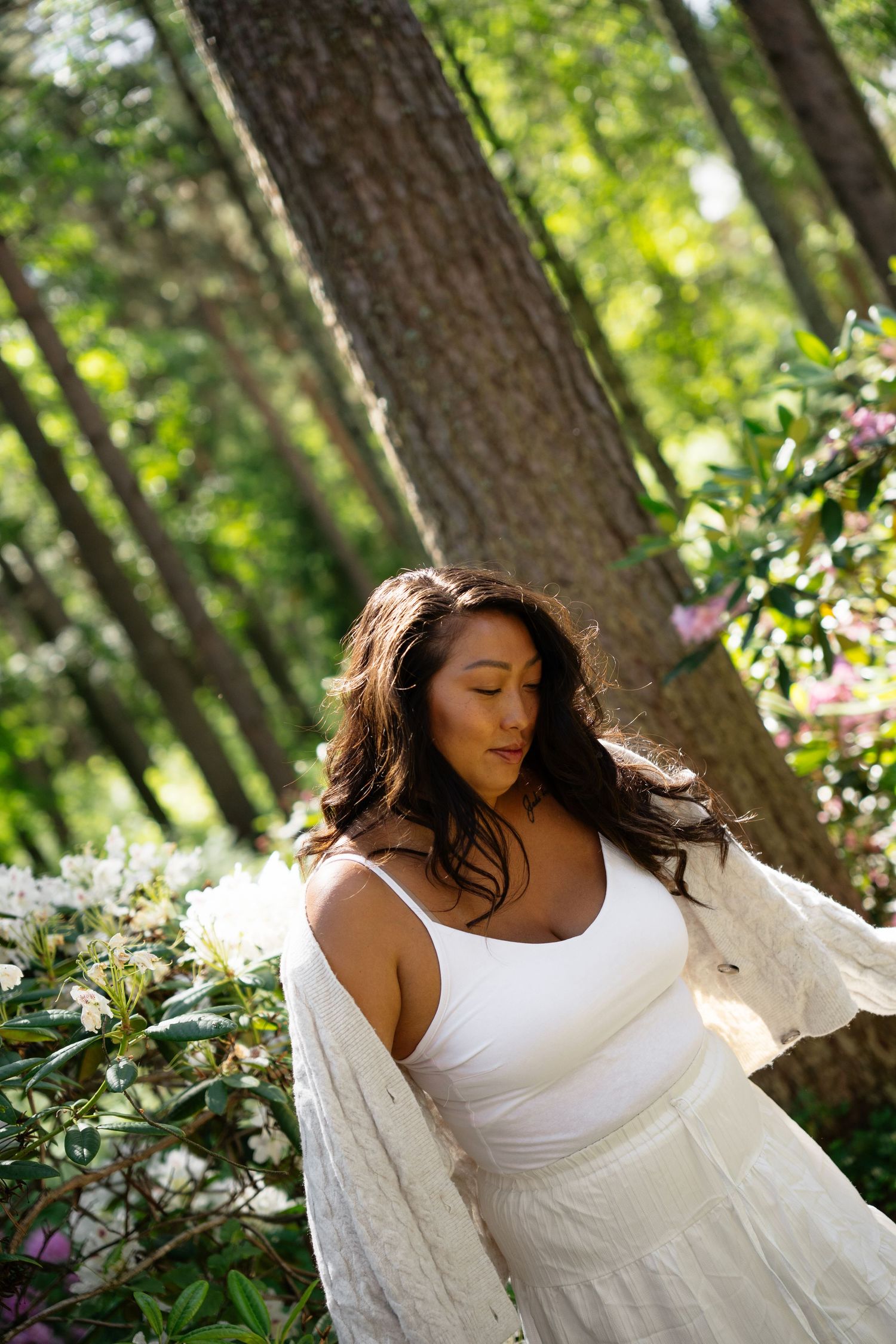 Woman in white dress standing among blooming flowers in a sunlit forest setting.
