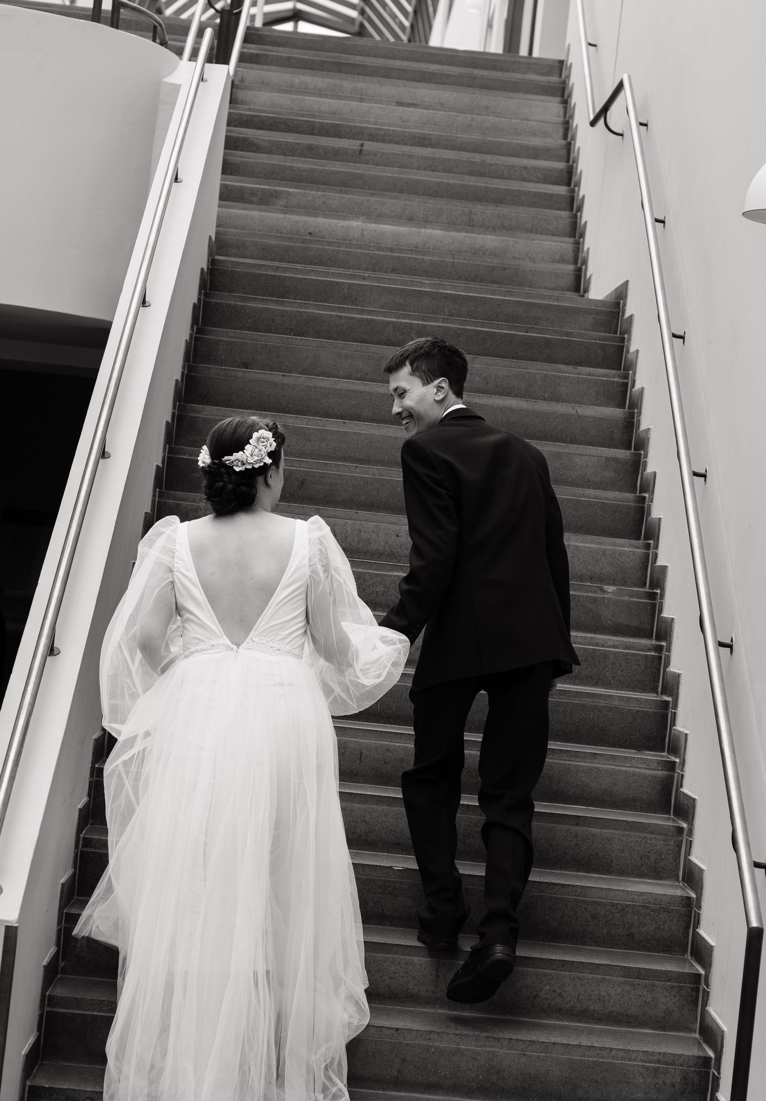 Black and white photo of a bride and groom holding hands walking up stairs, bride in elegant backless gown.