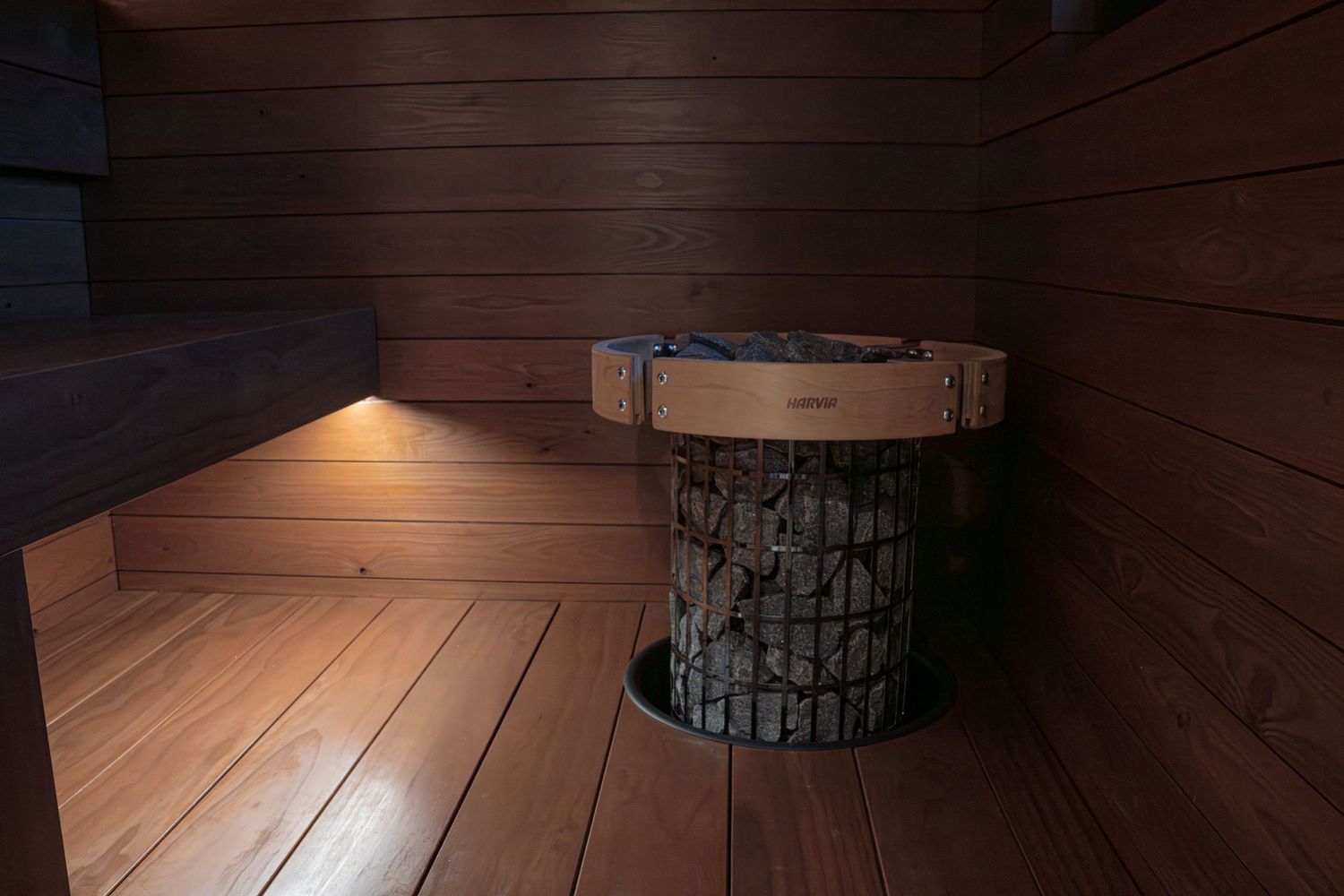 Wooden sauna interior with a round stone heater, warm lighting, and cedar wood paneling on walls and floor.