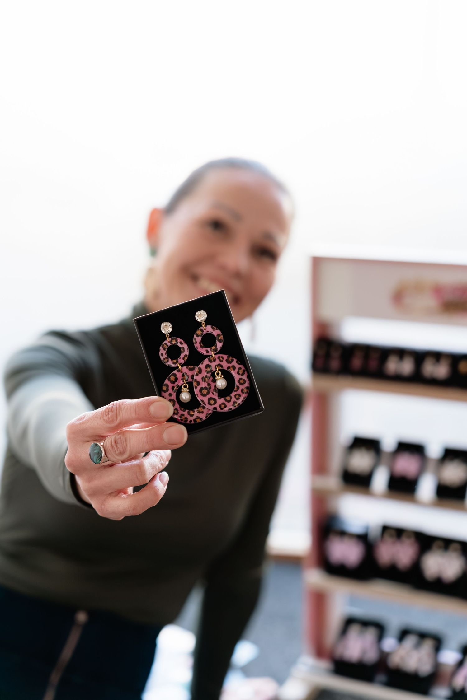 Woman smiling and holding pink leopard print drop earrings on black display card at jewelry stand.