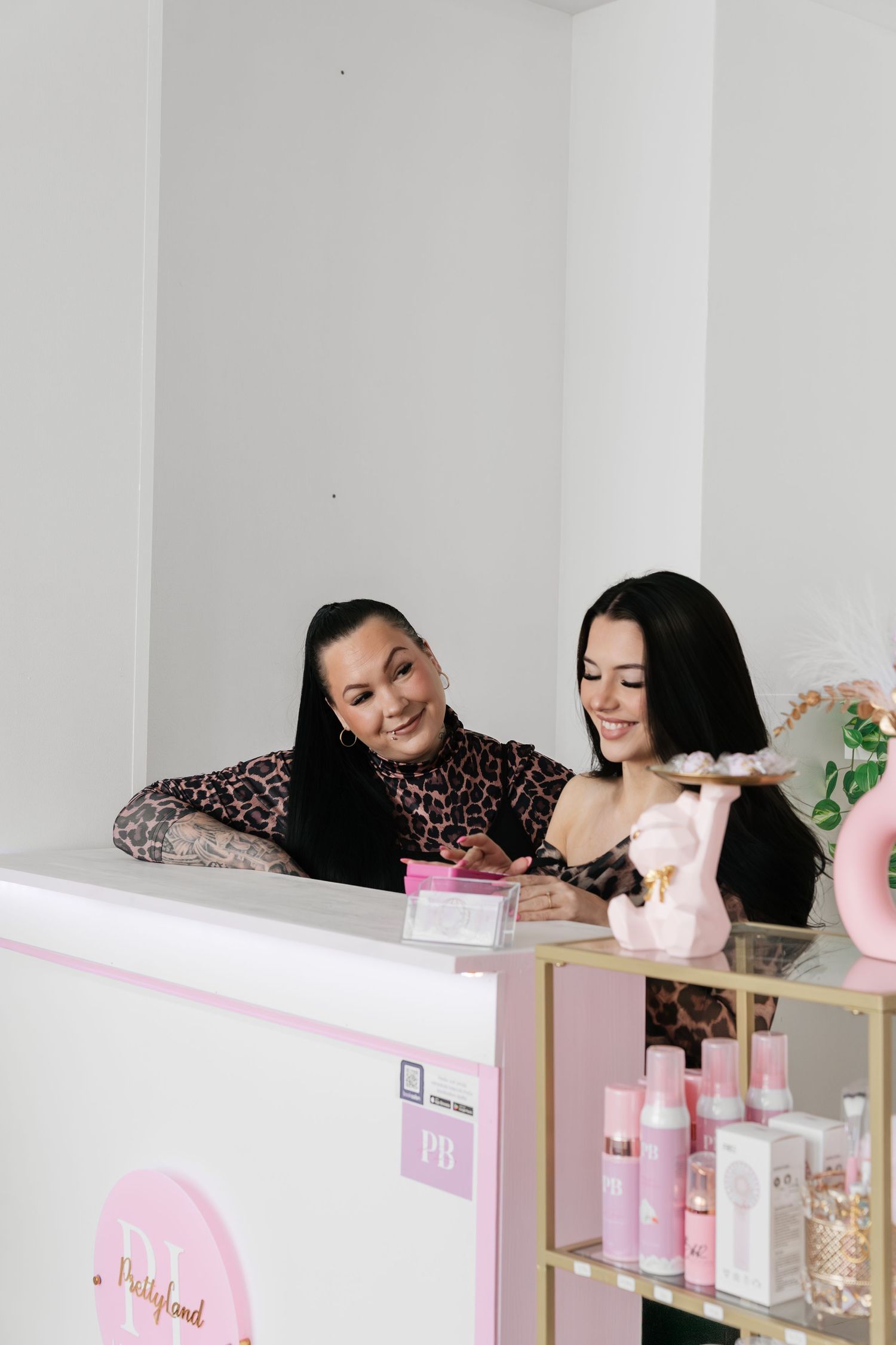 Two dark-haired women smiling behind a pink beauty salon reception desk with products displayed.