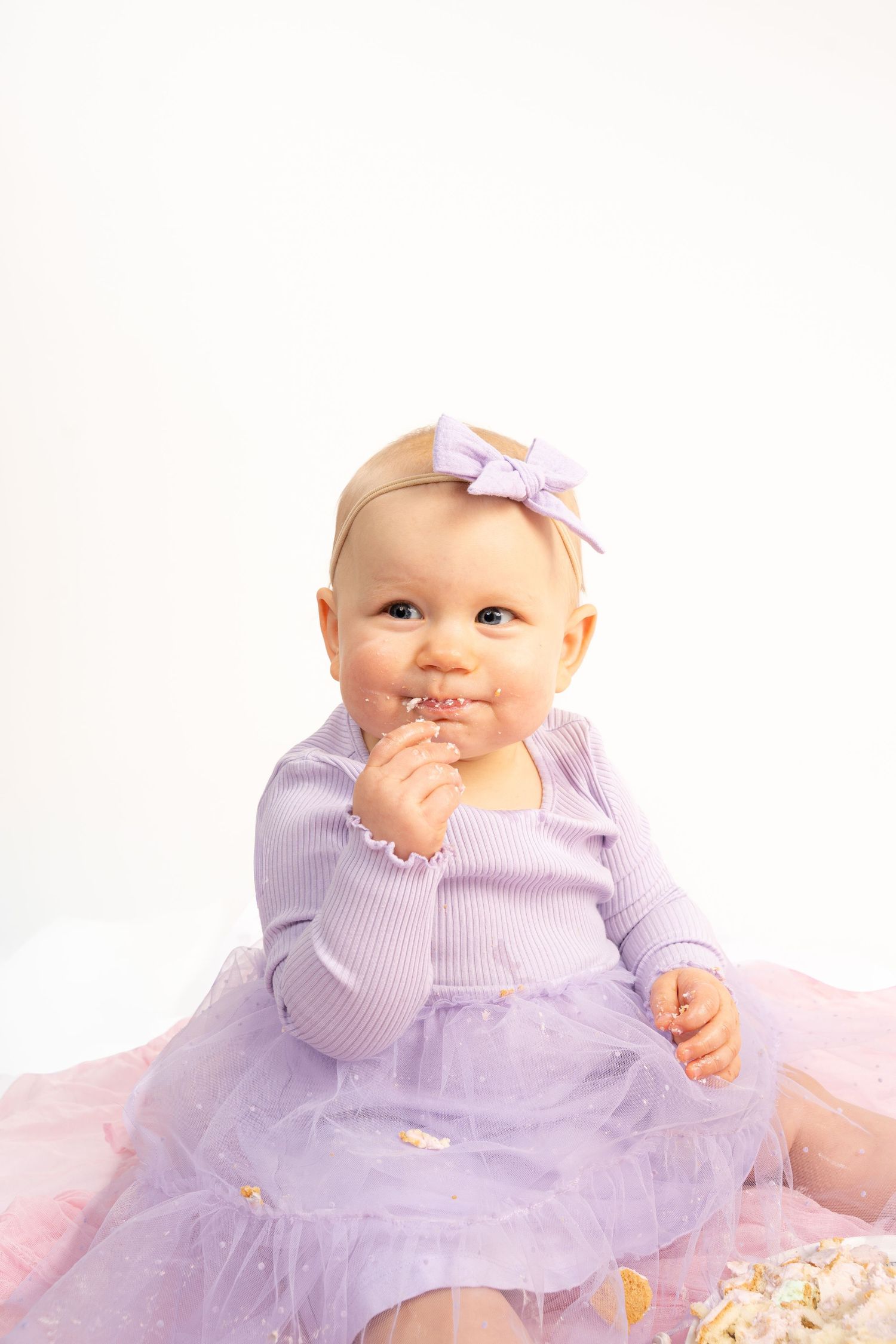Smiling baby girl in purple tutu dress and bow headband sitting on white background, fingers in mouth.