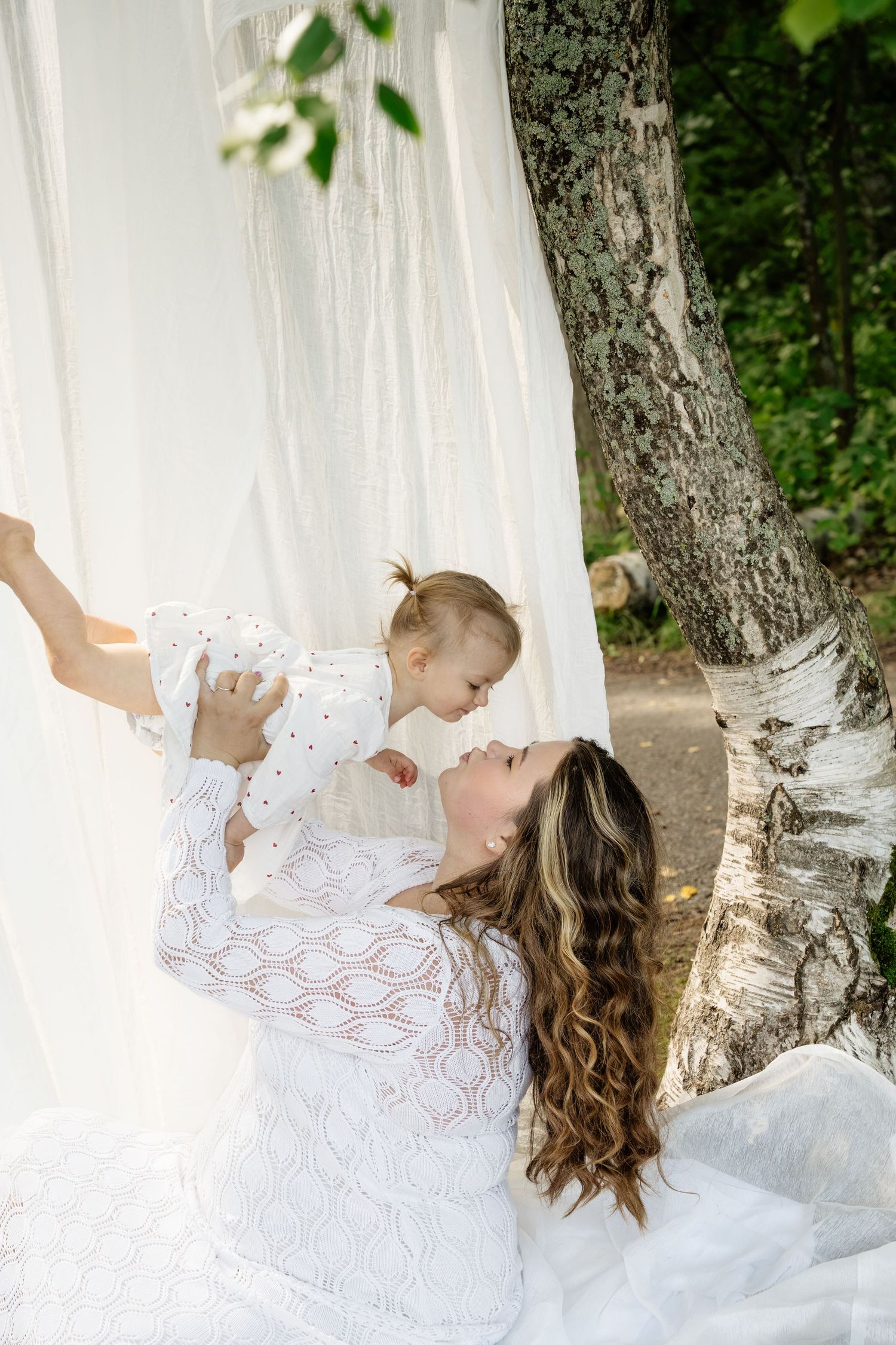 Mother in white lace dress lifts smiling baby outdoors near birch tree with flowing white curtains.