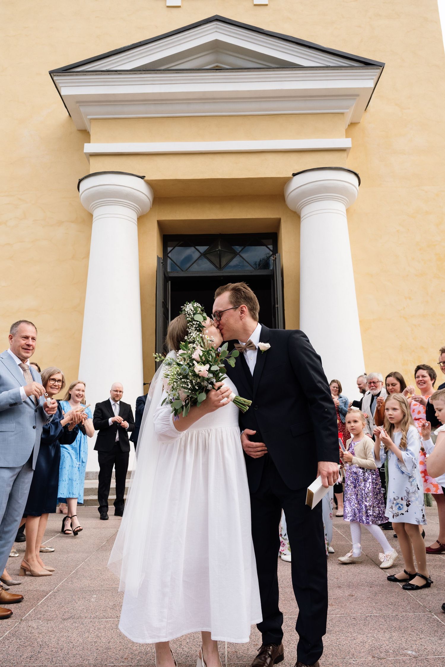 Newlyweds share a kiss outside a yellow church with white columns, surrounded by cheering wedding guests.