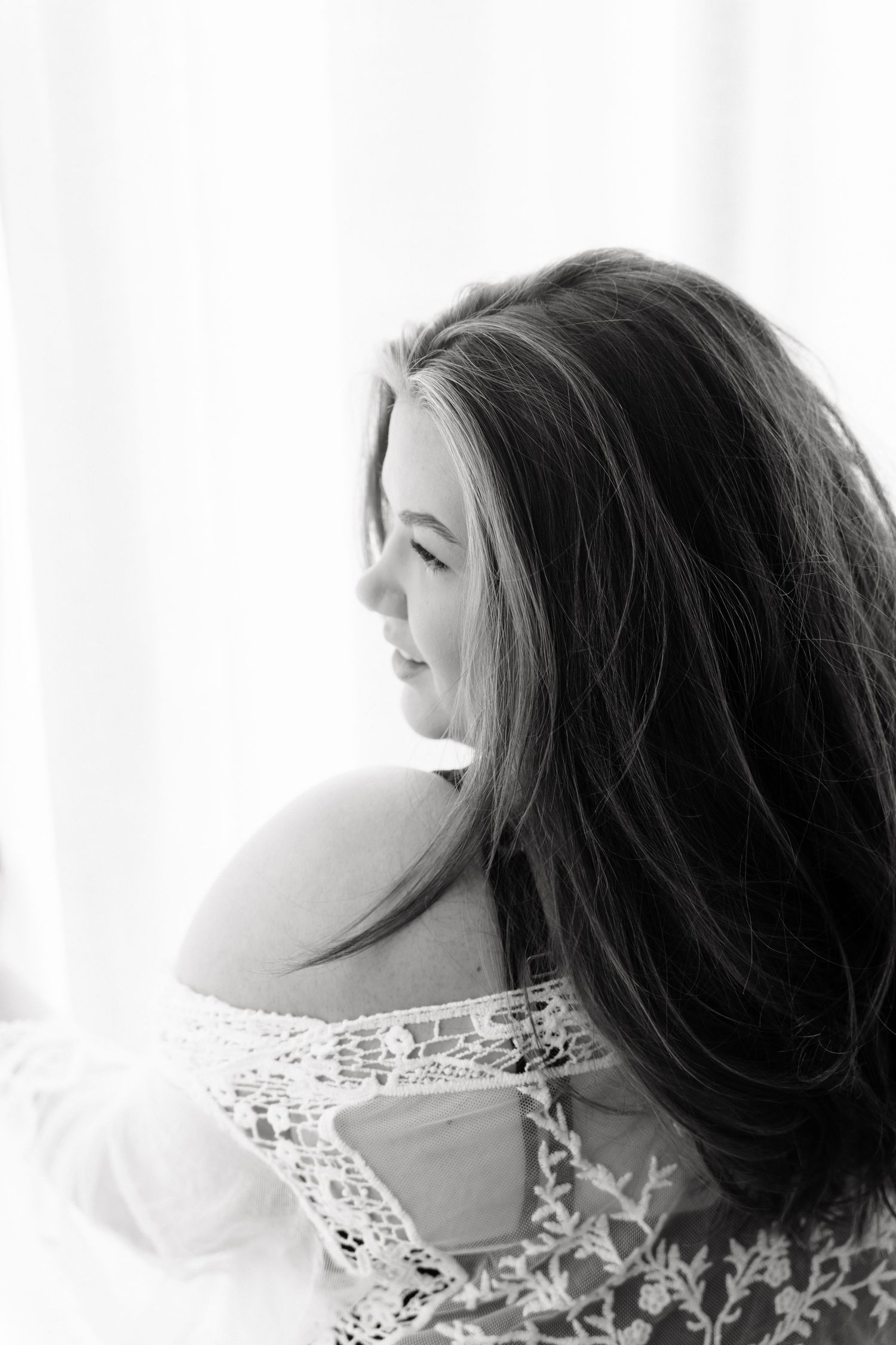 Black and white portrait of a woman with long dark hair wearing lace dress, looking sideways near bright window.