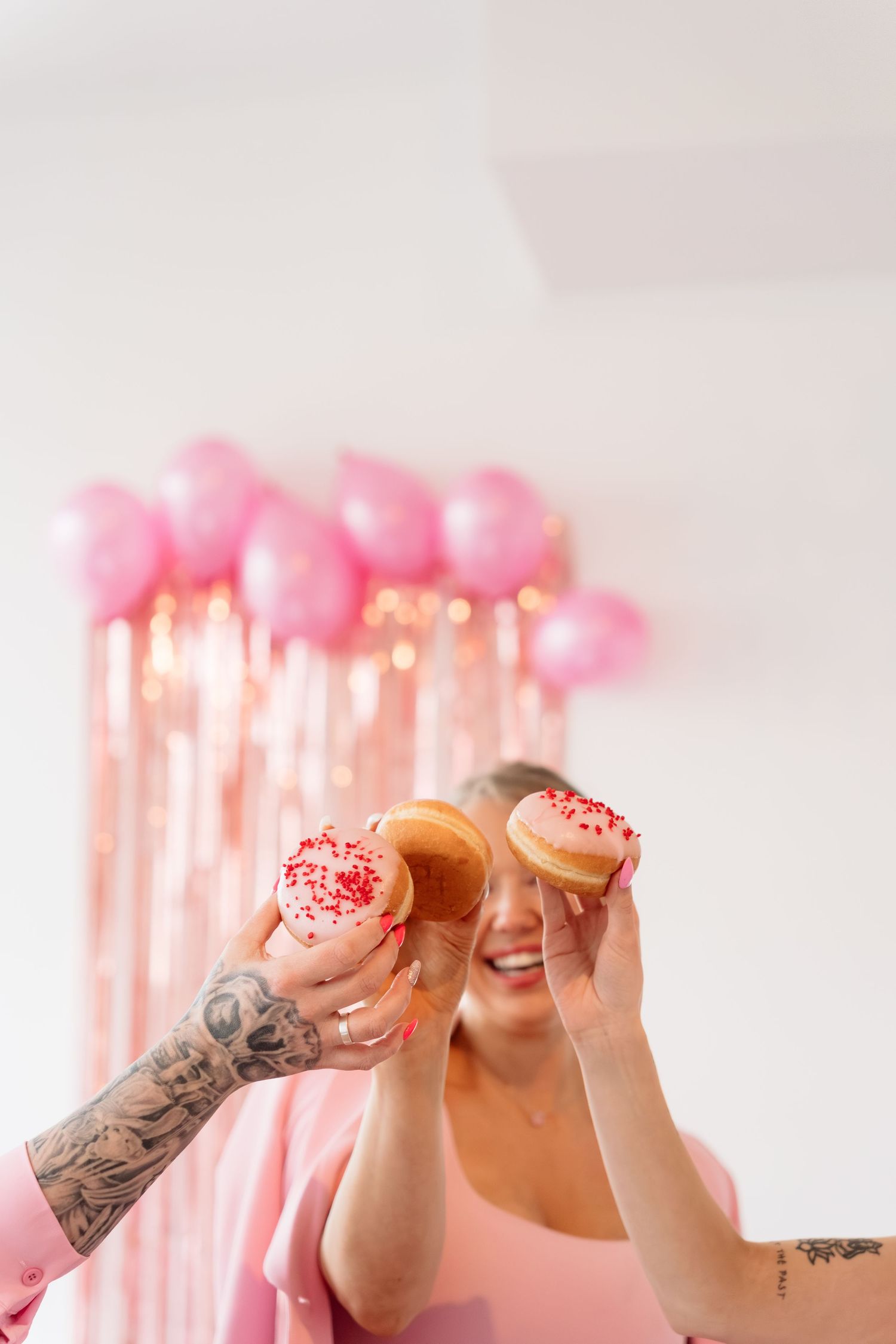 Happy woman with tattoos holding pink frosted donuts in front of pink balloon party decorations.