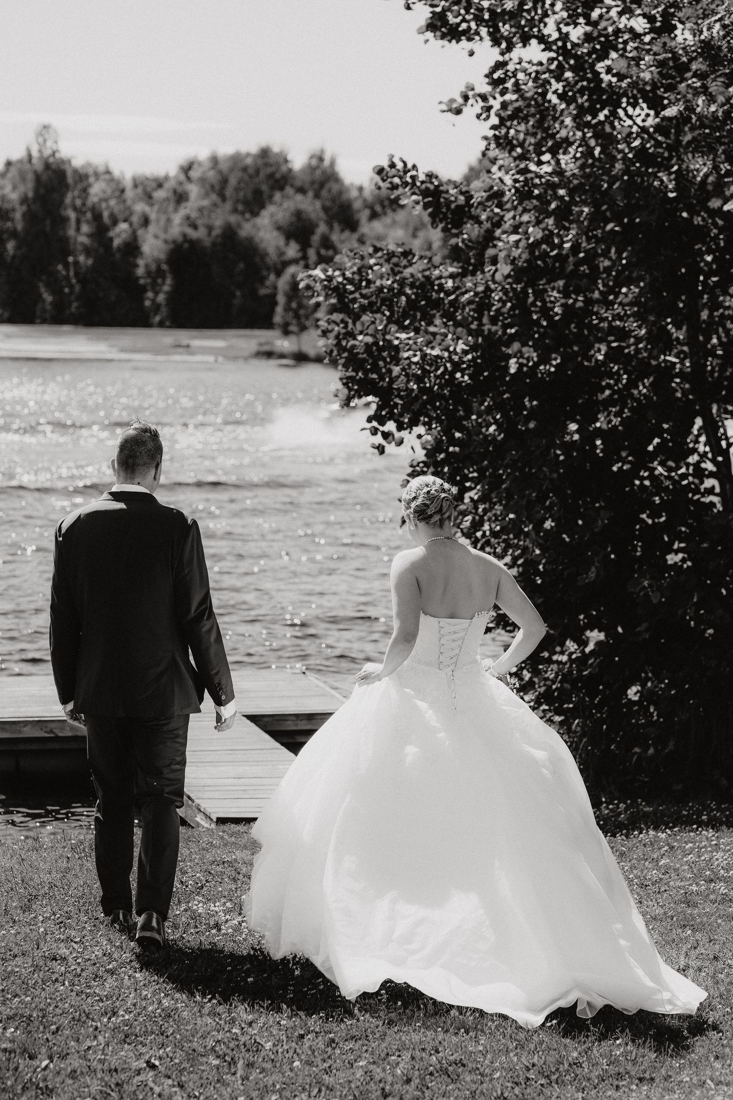 Black and white photo of bride and groom walking toward a sunlit lake, viewed from behind.