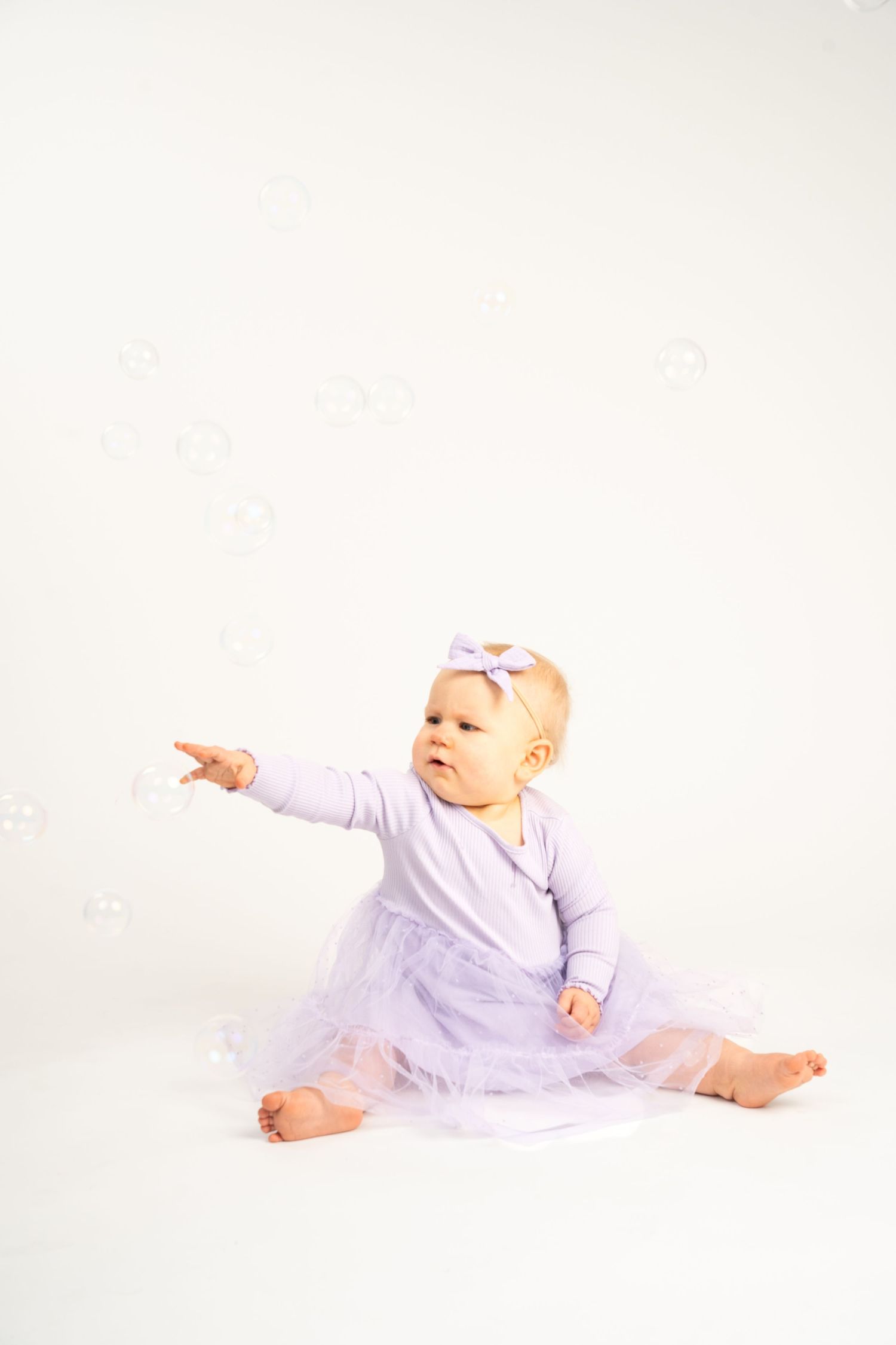 A baby girl in a purple tutu dress reaches for floating soap bubbles on a white background.