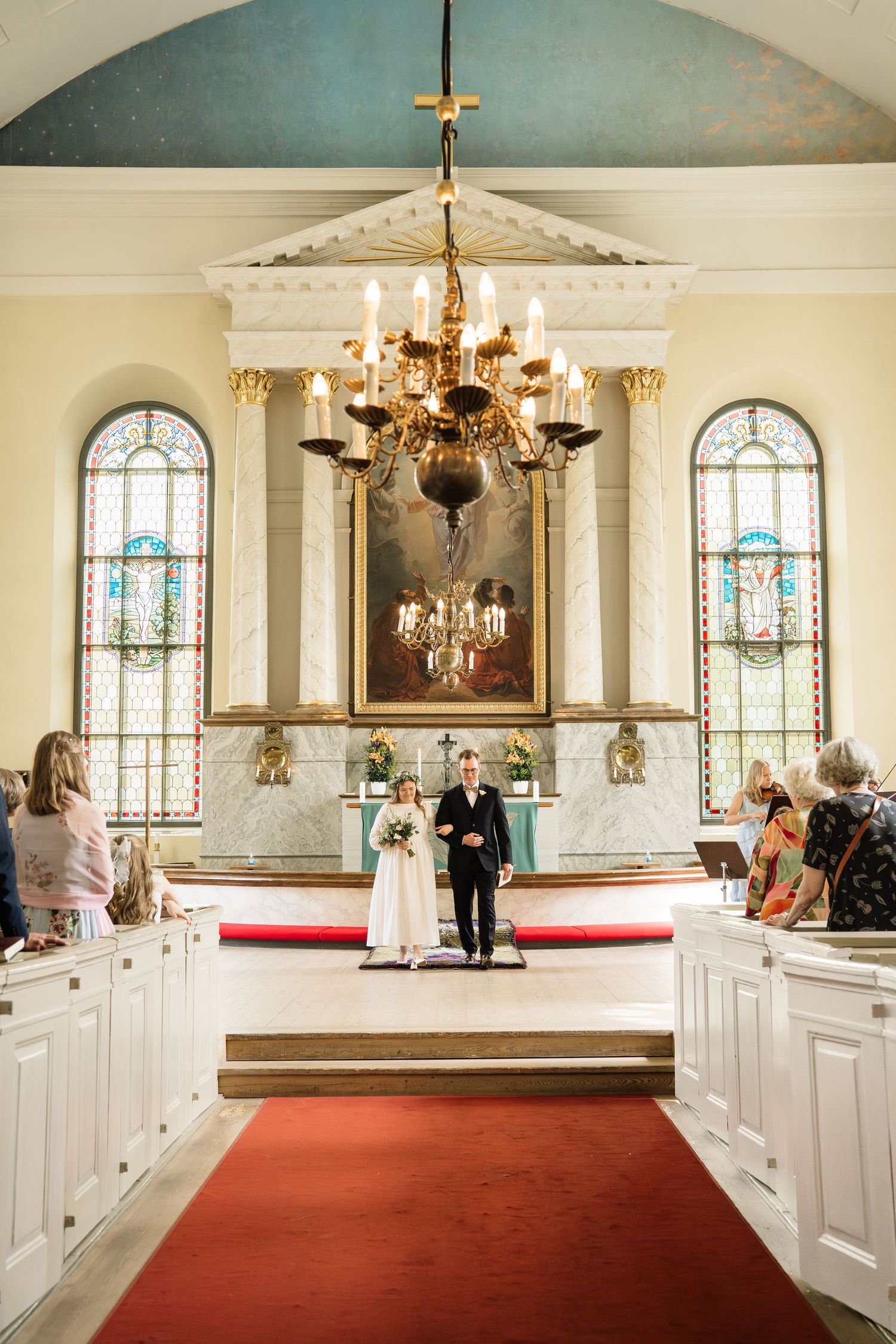 Bride and groom stand at the altar of an elegant church with stained glass windows and a gold chandelier.