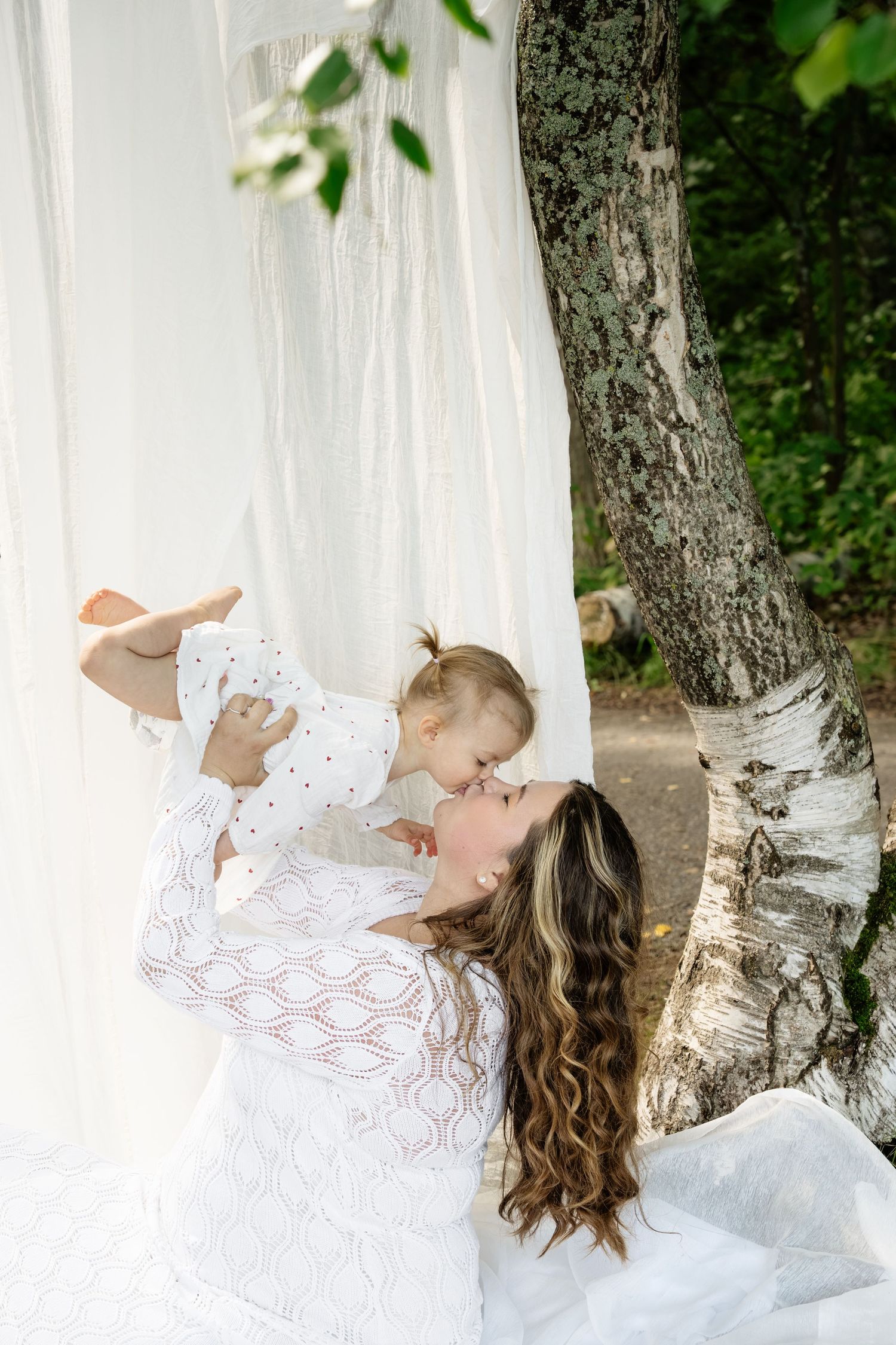 Mother in white lace dress lifting and kissing her baby outdoors near birch tree with flowing white curtains.