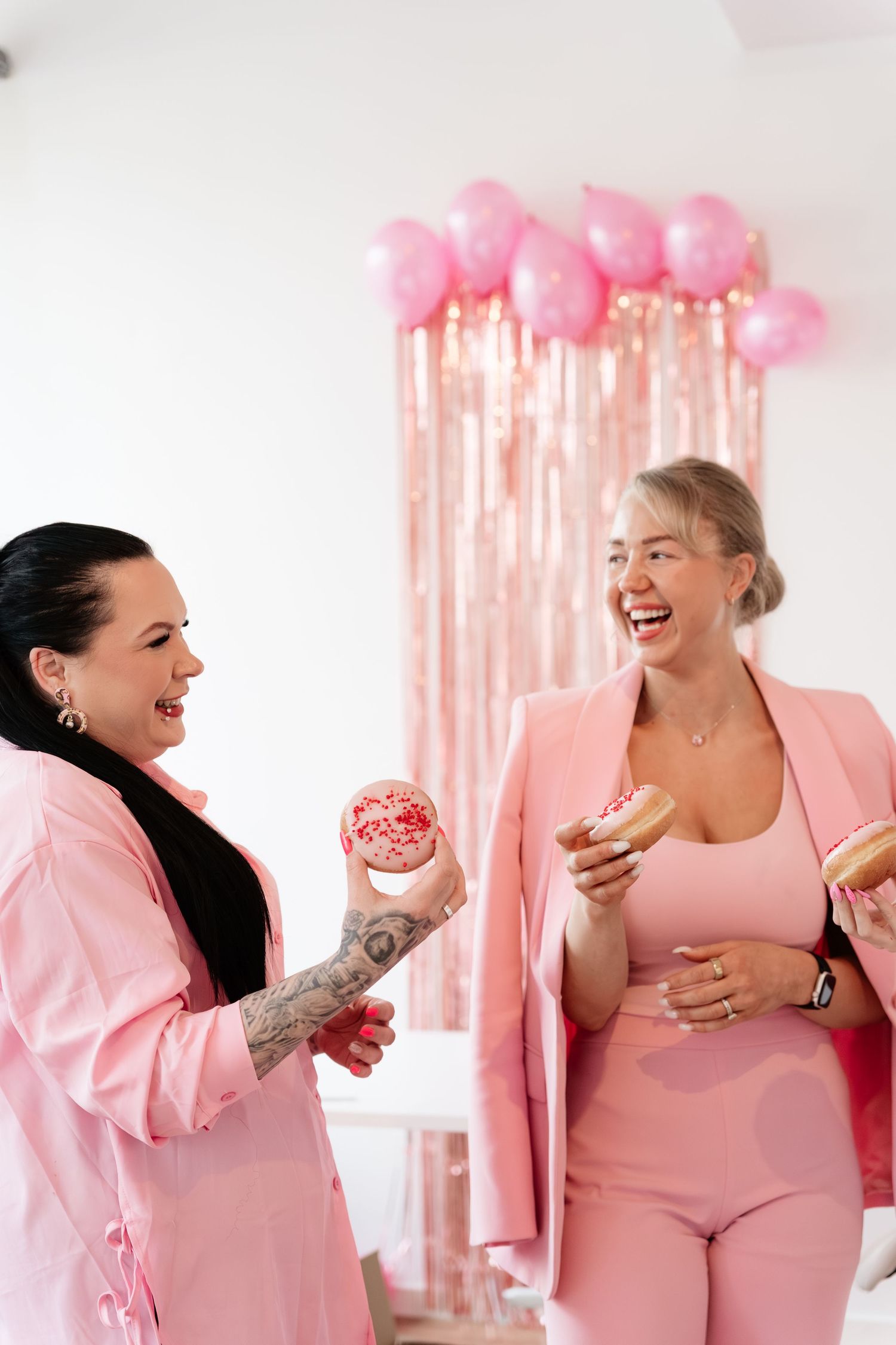 Two women in pink outfits laughing while holding pink frosted donuts at party celebration.