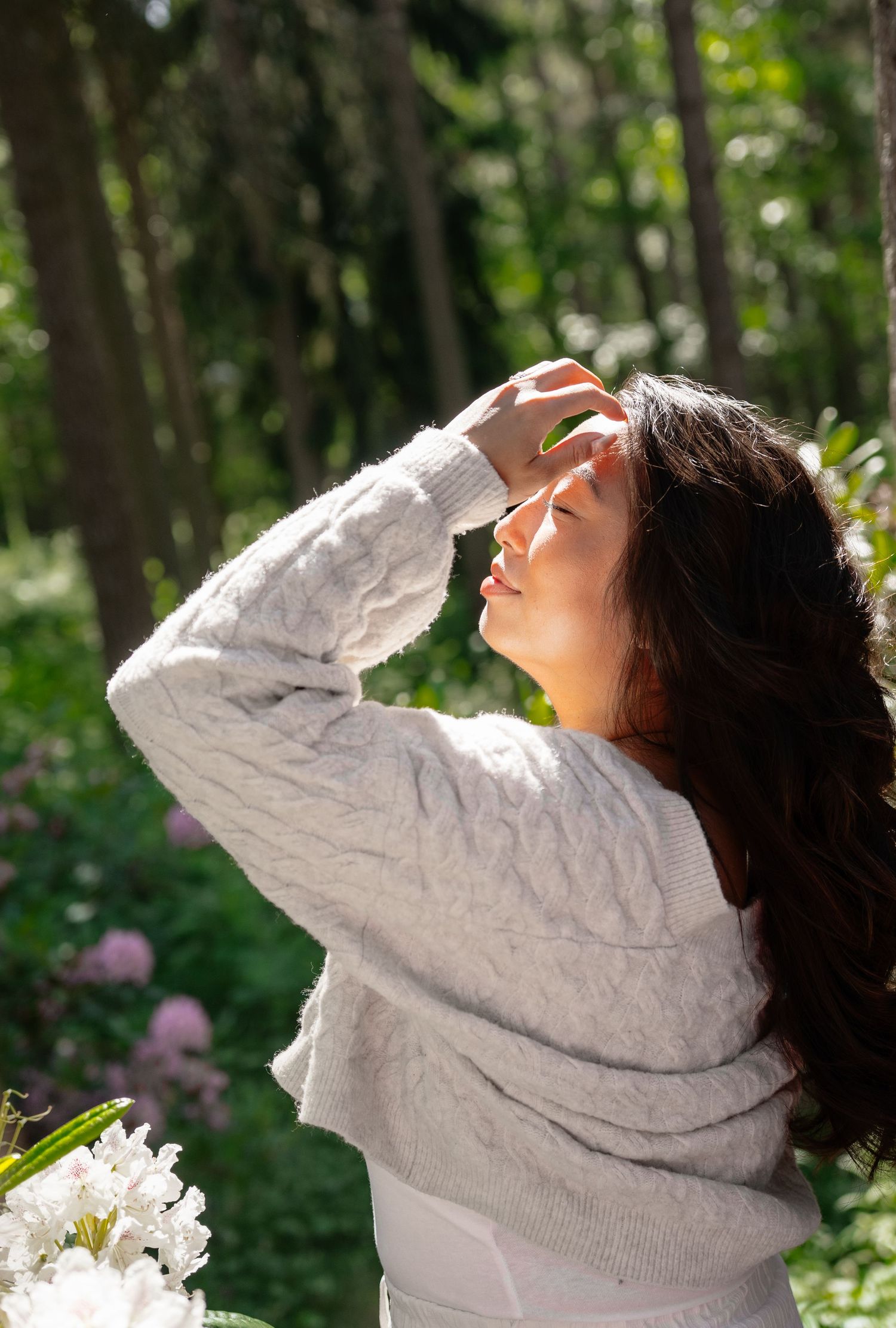 Young woman in white cable-knit sweater shielding her eyes from sunlight in a lush green forest.