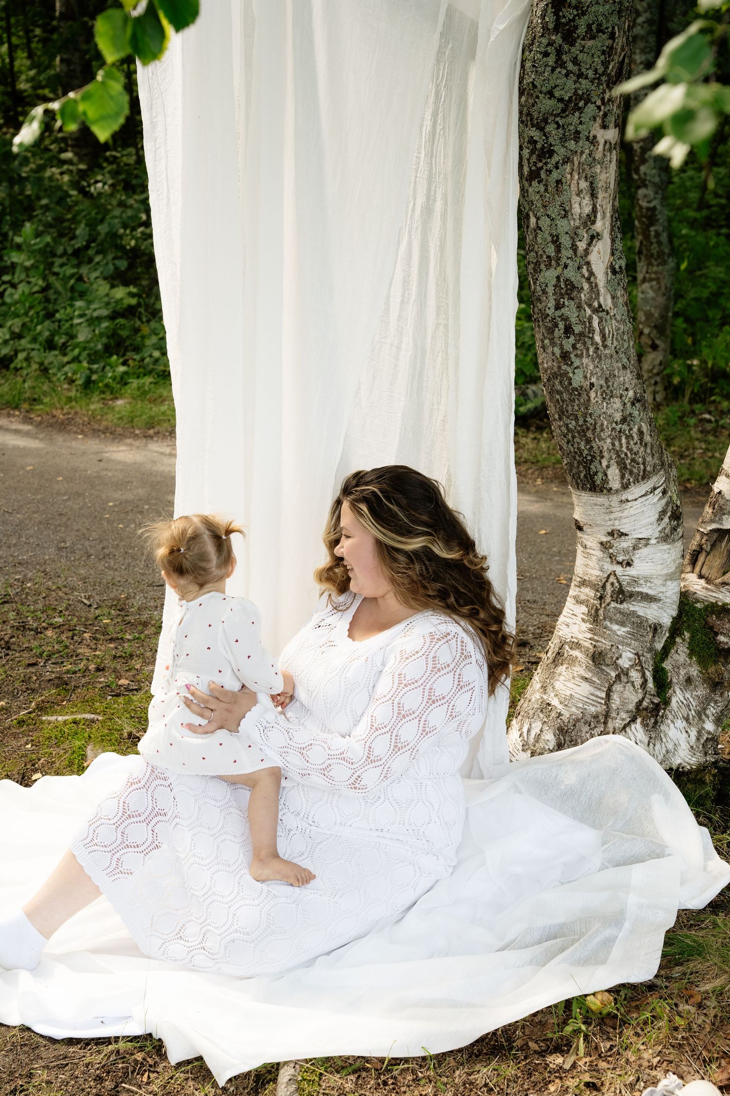 A pregnant woman in a white lace dress sits on a blanket outdoors with her toddler daughter near birch trees.