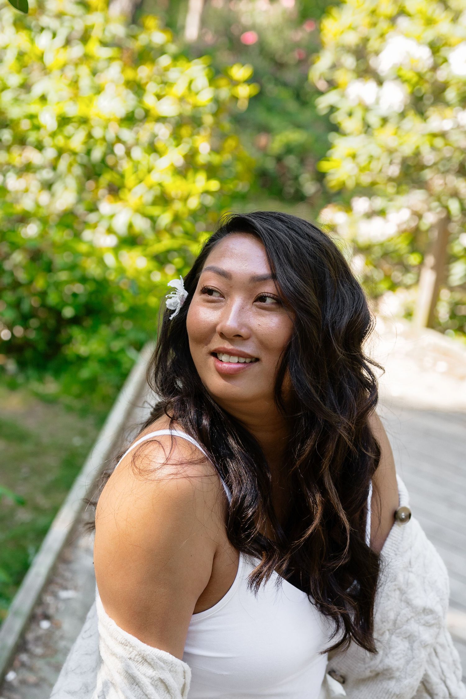 Smiling Asian woman with dark wavy hair and white flower accessory wearing a white top in a sunny green park.