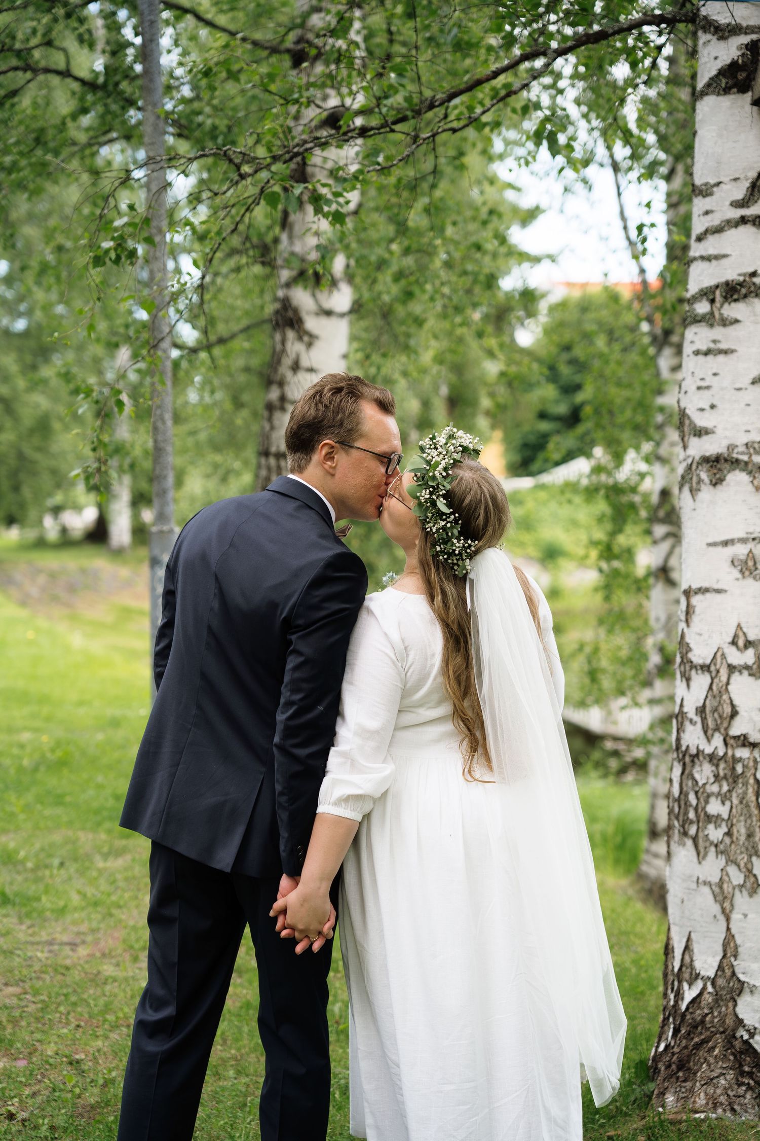 Bride and groom kissing under birch trees, bride wearing floral crown and veil, groom in dark suit, outdoor wedding.