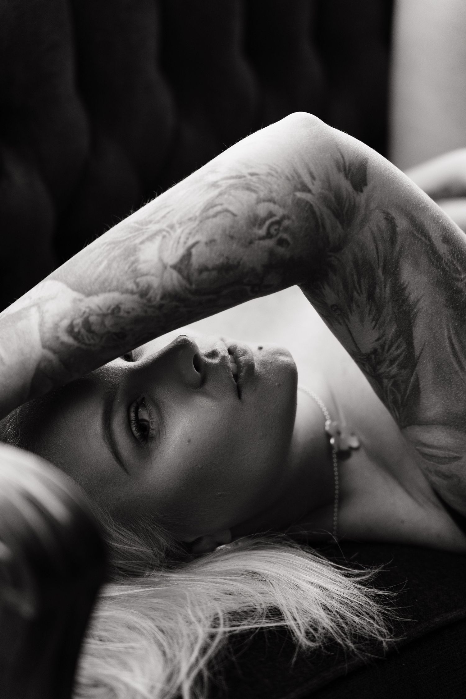 Black and white boudoir portrait of blonde woman with tattoed arm resting above her head.
