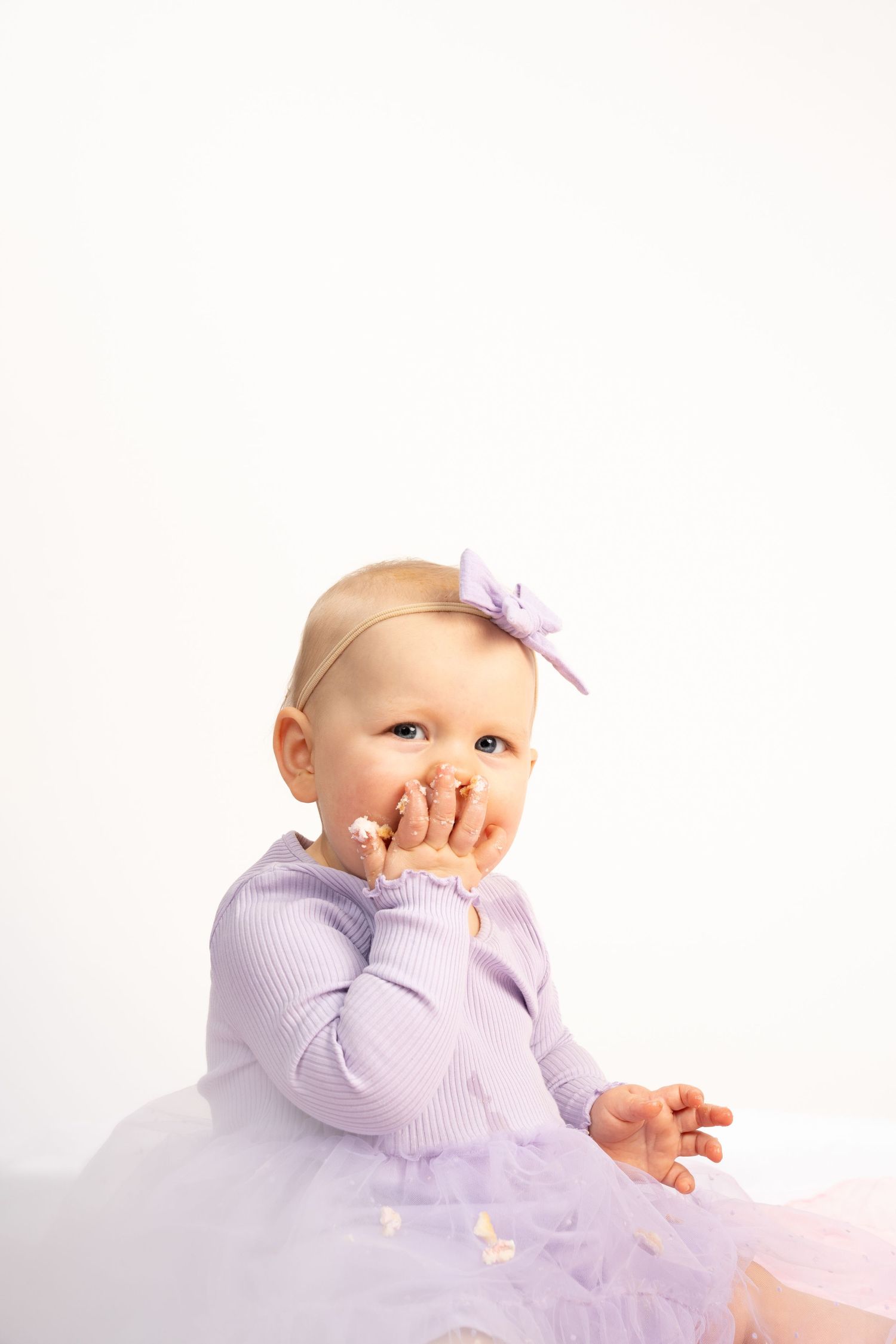 A cute baby girl in a lavender tutu dress and bow headband sits on a white background, covering her mouth with her hand.