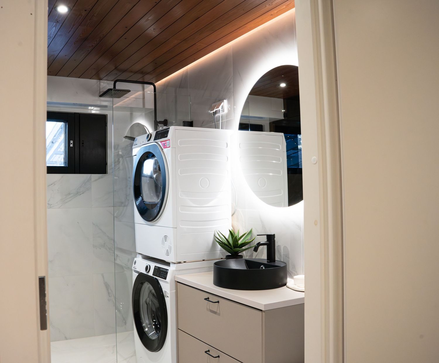 Modern bathroom with stacked washer-dryer, marble walls, round backlit mirror, black vessel sink vanity, and walk-in shower.
