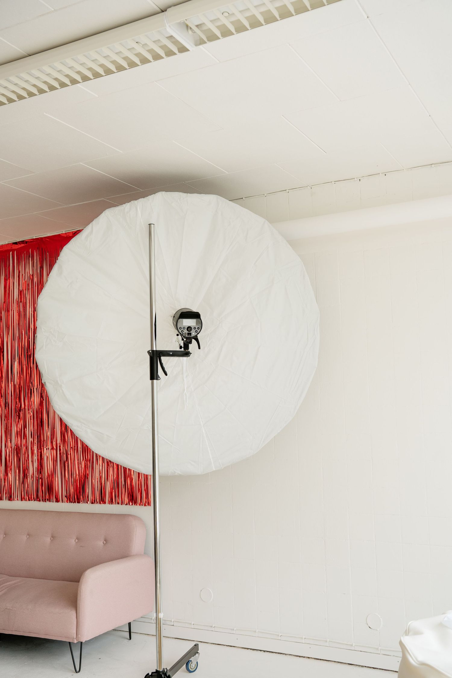 Large white photography umbrella on a stand in a studio with a pink sofa and red backdrop.