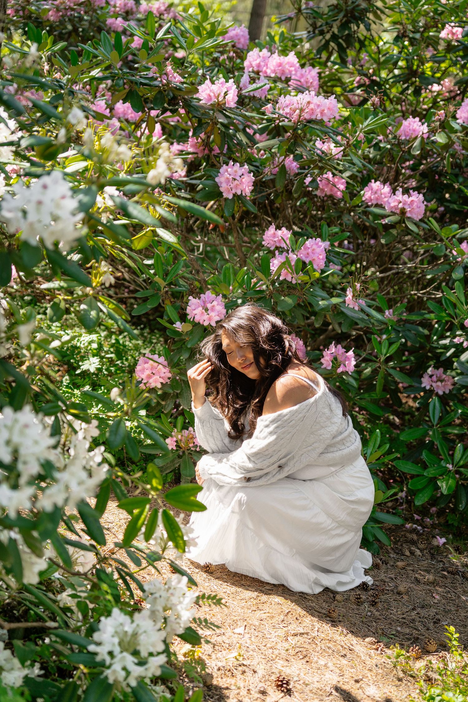 Woman in white off-shoulder dress crouching among pink and white rhododendron blooms in a garden.