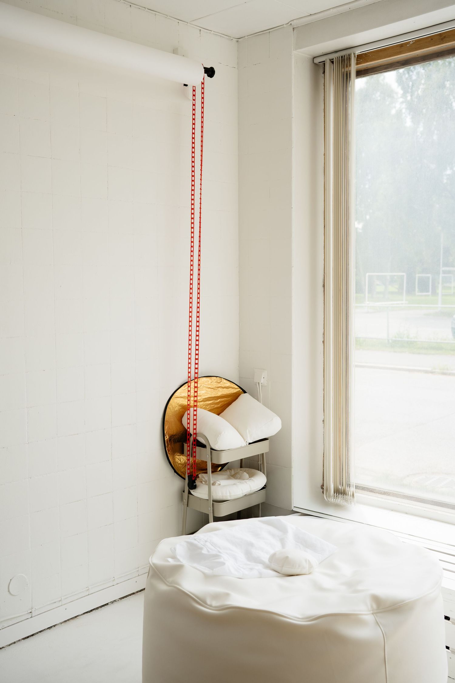 Minimalist white room corner with a massage table, round mirror, metal cart, and red dotted string near a large window.