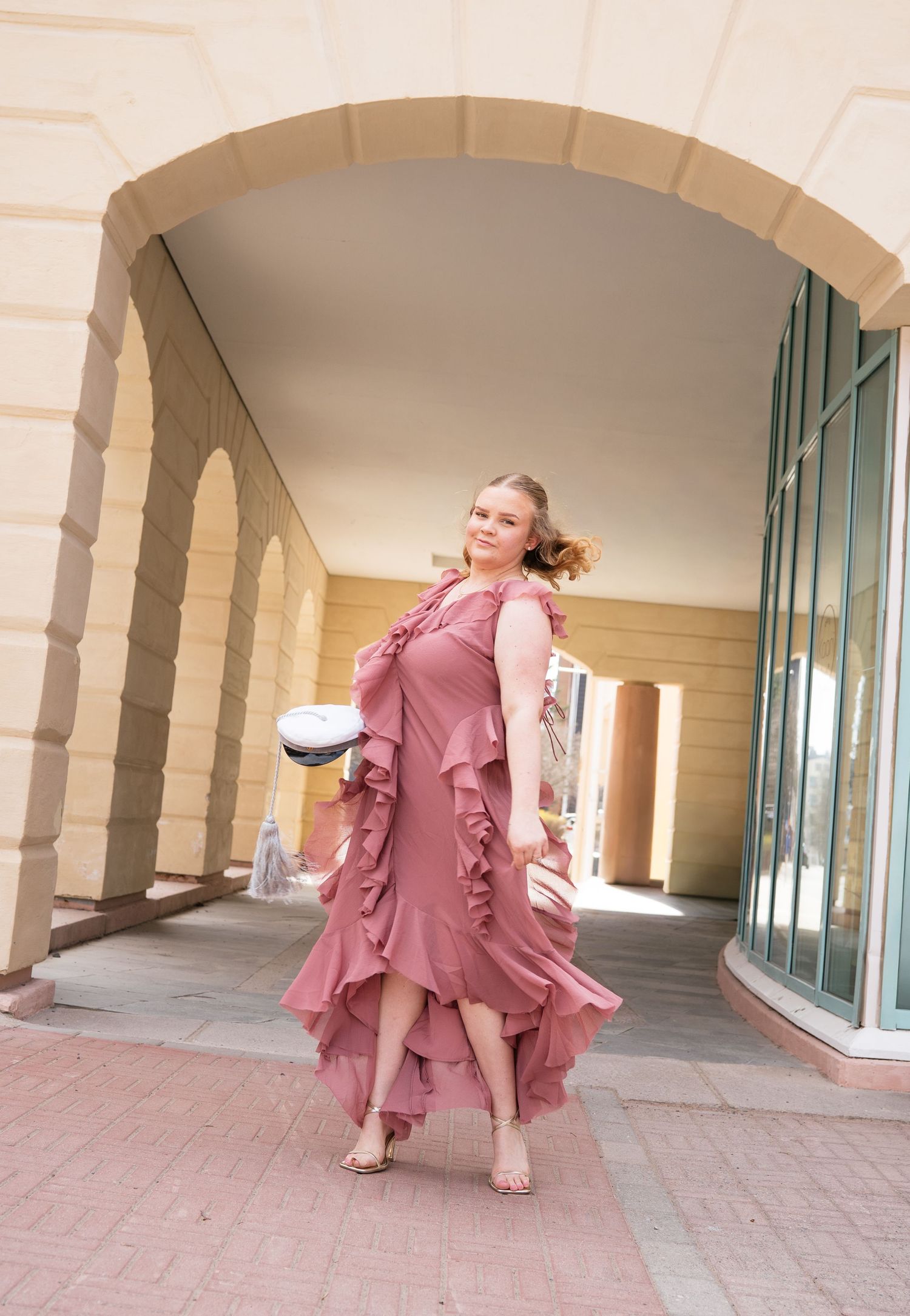 Young woman in a pink ruffled maxi dress holding a graduation cap under an elegant arched colonnade.