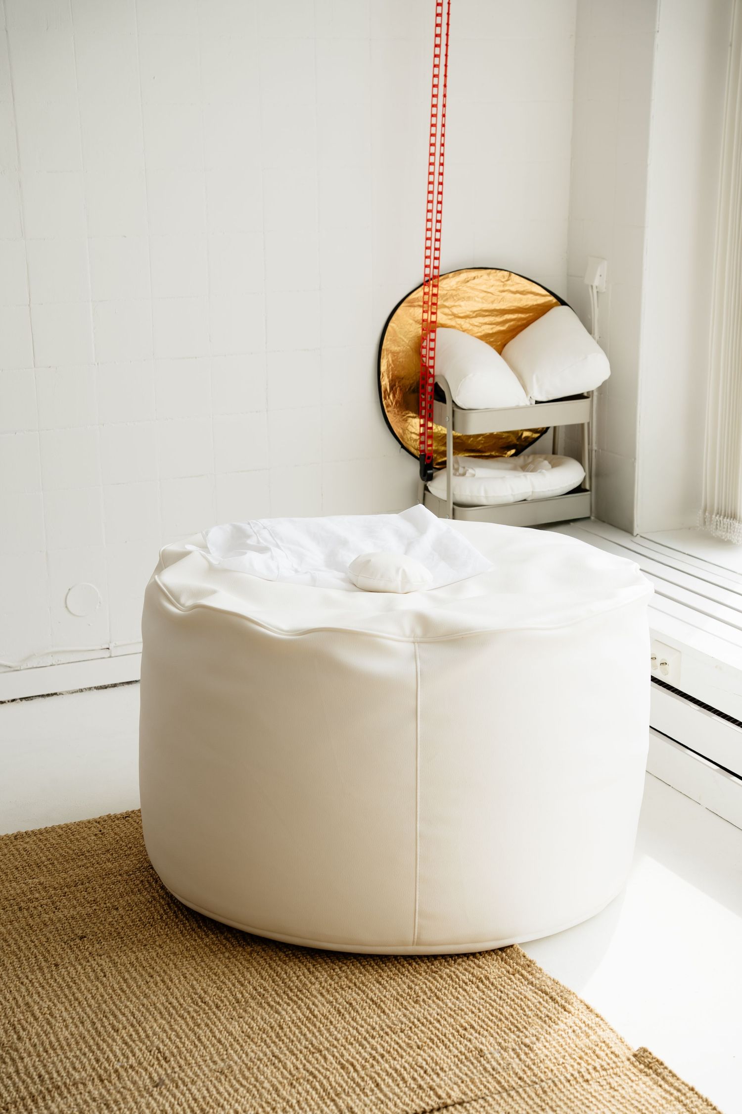 White leather bean bag chair on jute rug in minimalist white room with gold mirror and chrome chair.