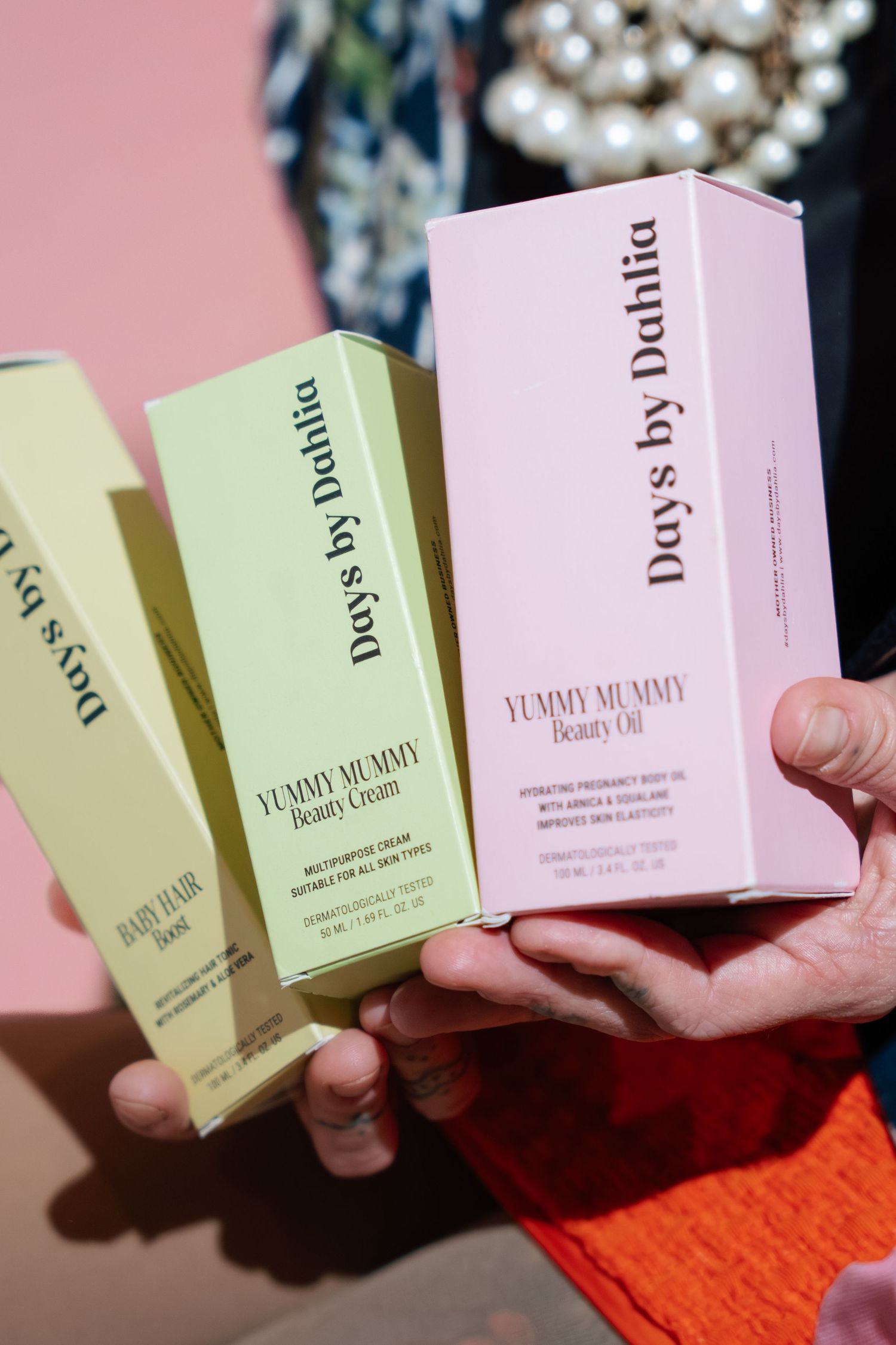 Hands holding three Days by Dahlia Yummy Mummy beauty product boxes against a pink background.