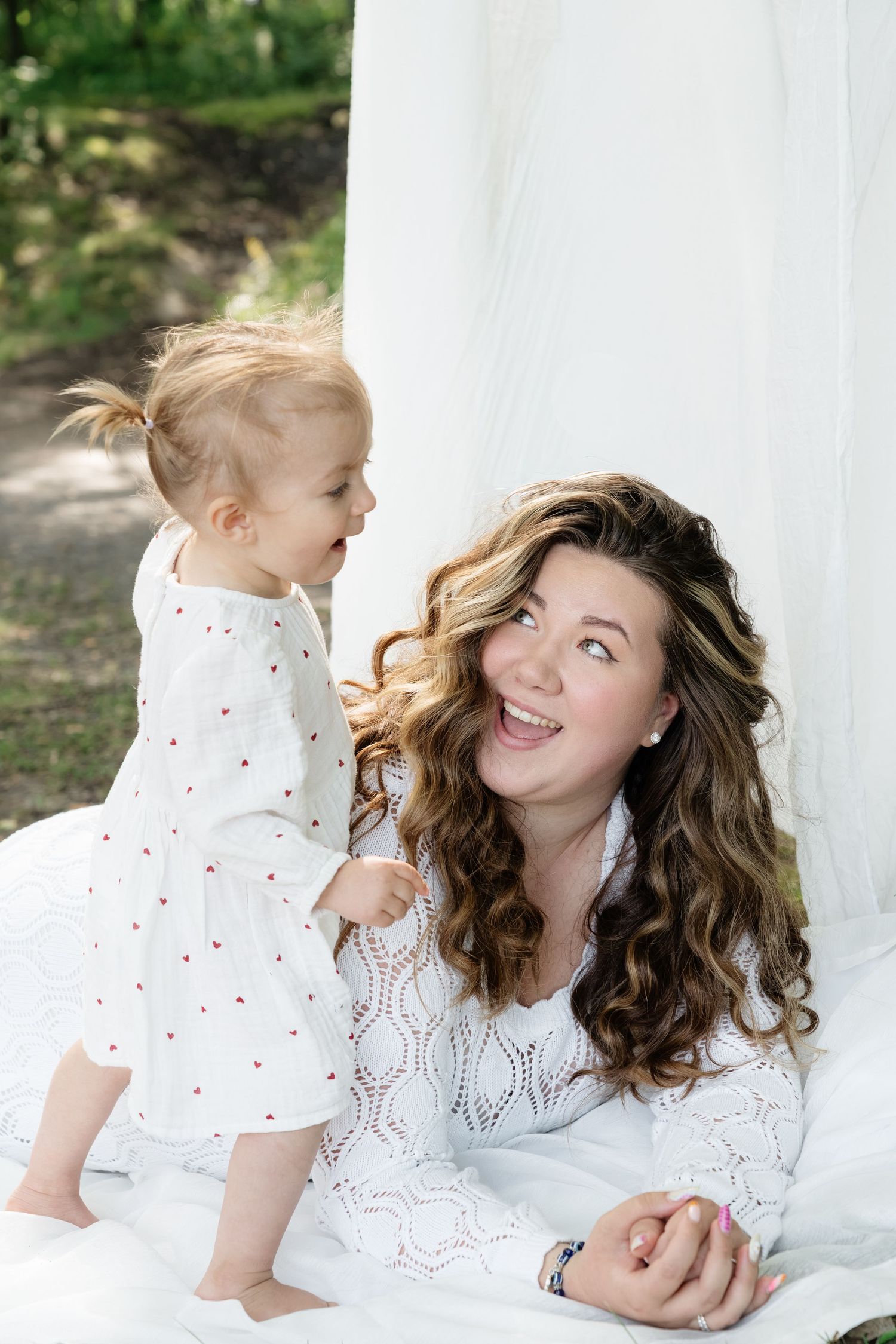 Happy young mother in white dress smiles at toddler girl in polka dot dress outdoors.