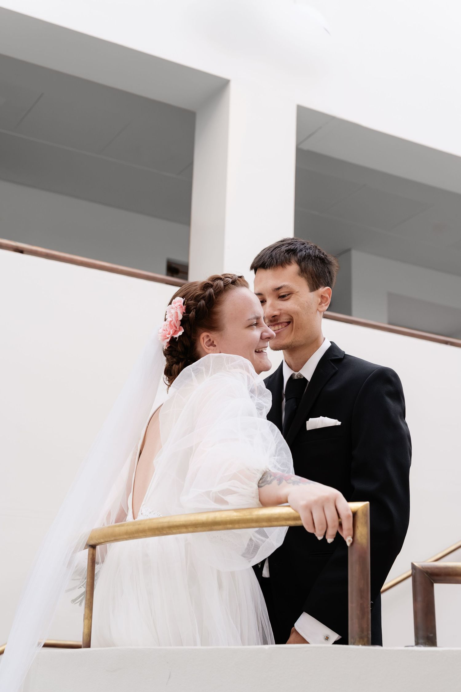 A smiling bride in a white gown and veil stands with a groom in a black suit on a staircase.