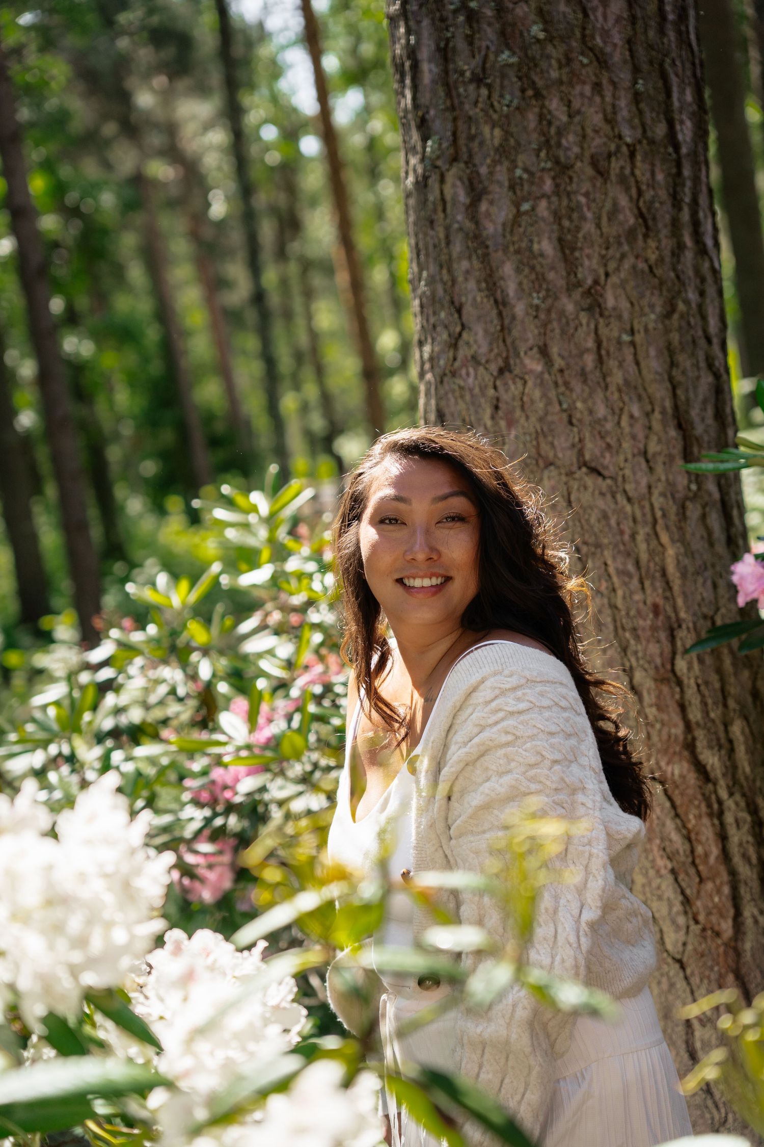 Smiling Asian woman in white cardigan leaning against tree trunk surrounded by blooming white flowers in sunny forest.