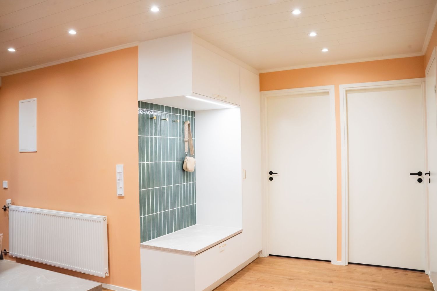 Modern entryway with peach walls, white built-in bench, teal tile accent, white doors, and wood flooring.
