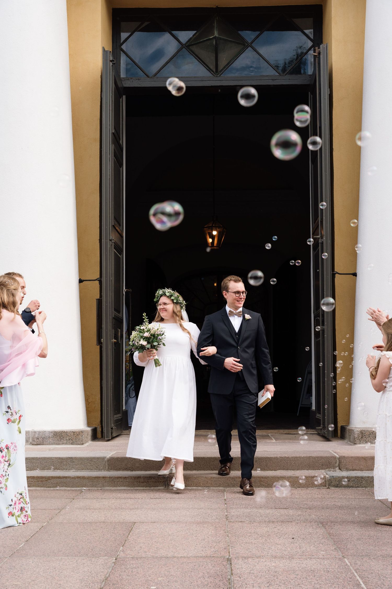 Bride and groom exit church doors surrounded by floating soap bubbles, guests celebrating on steps.