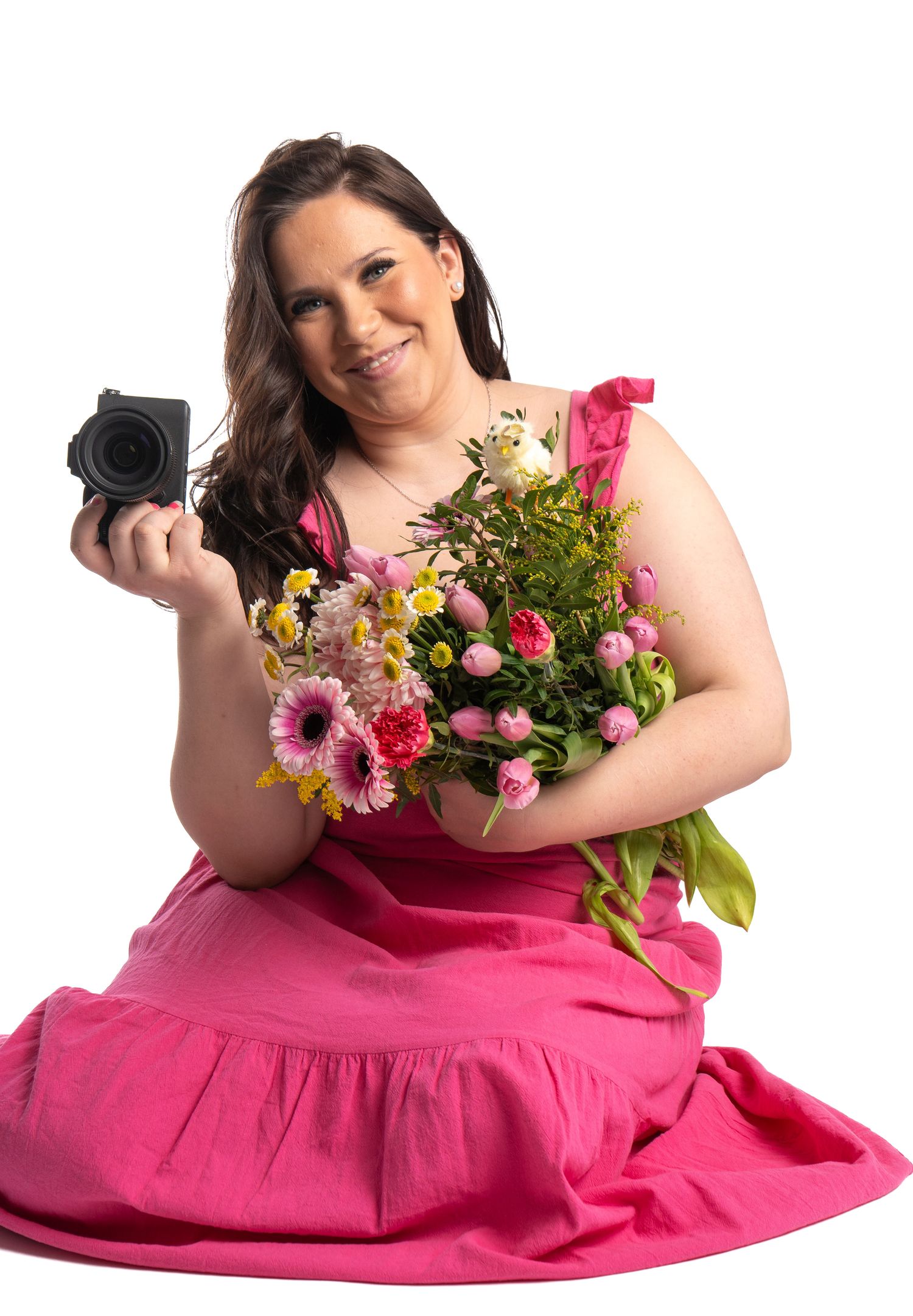 Smiling woman in pink dress holding camera and colorful flower bouquet on white background.