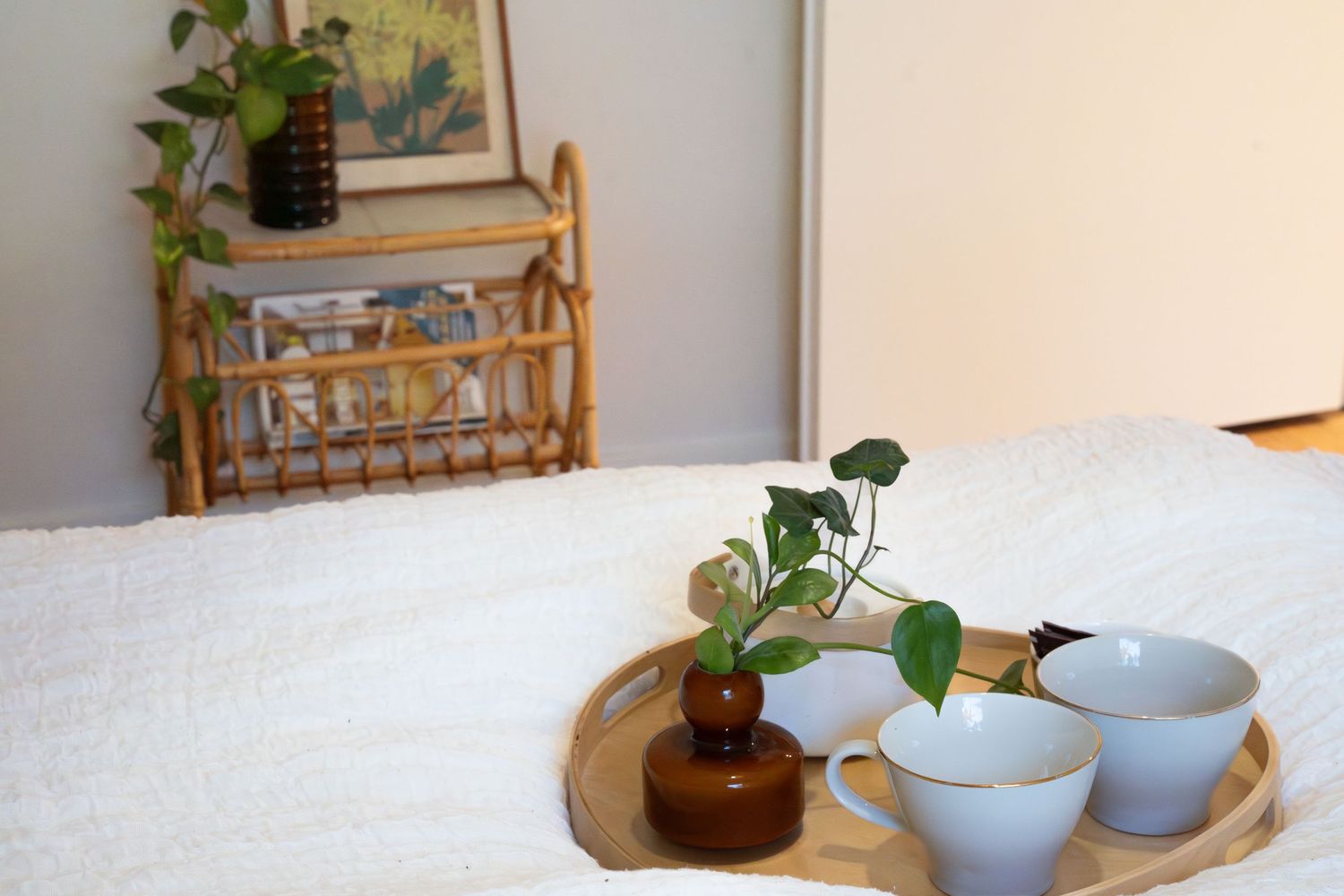 Wooden tray with two ceramic cups and a small plant vase on a white fluffy surface near a rattan shelf.