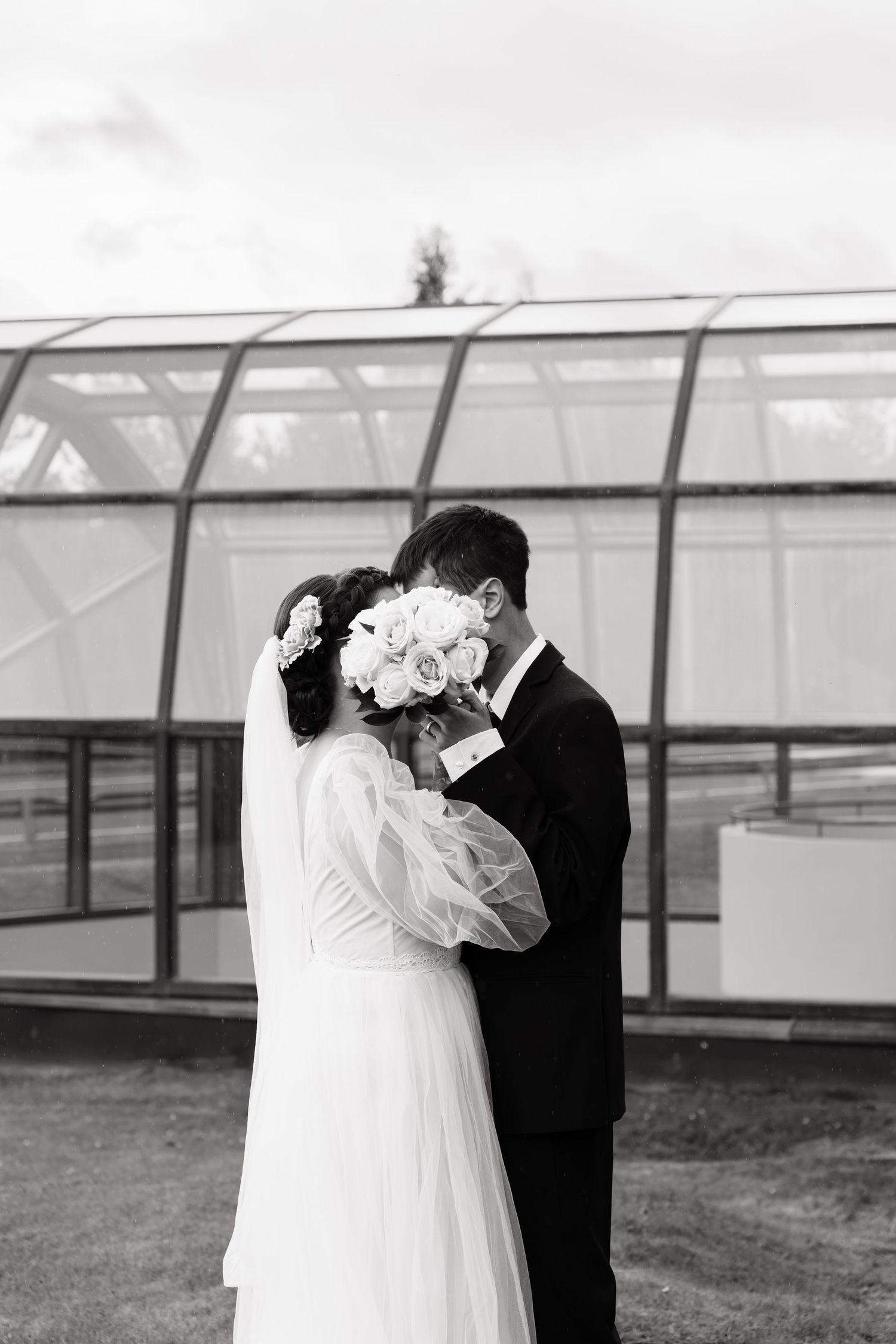 Black and white wedding photo of bride and groom kissing behind bridal bouquet outside greenhouse.