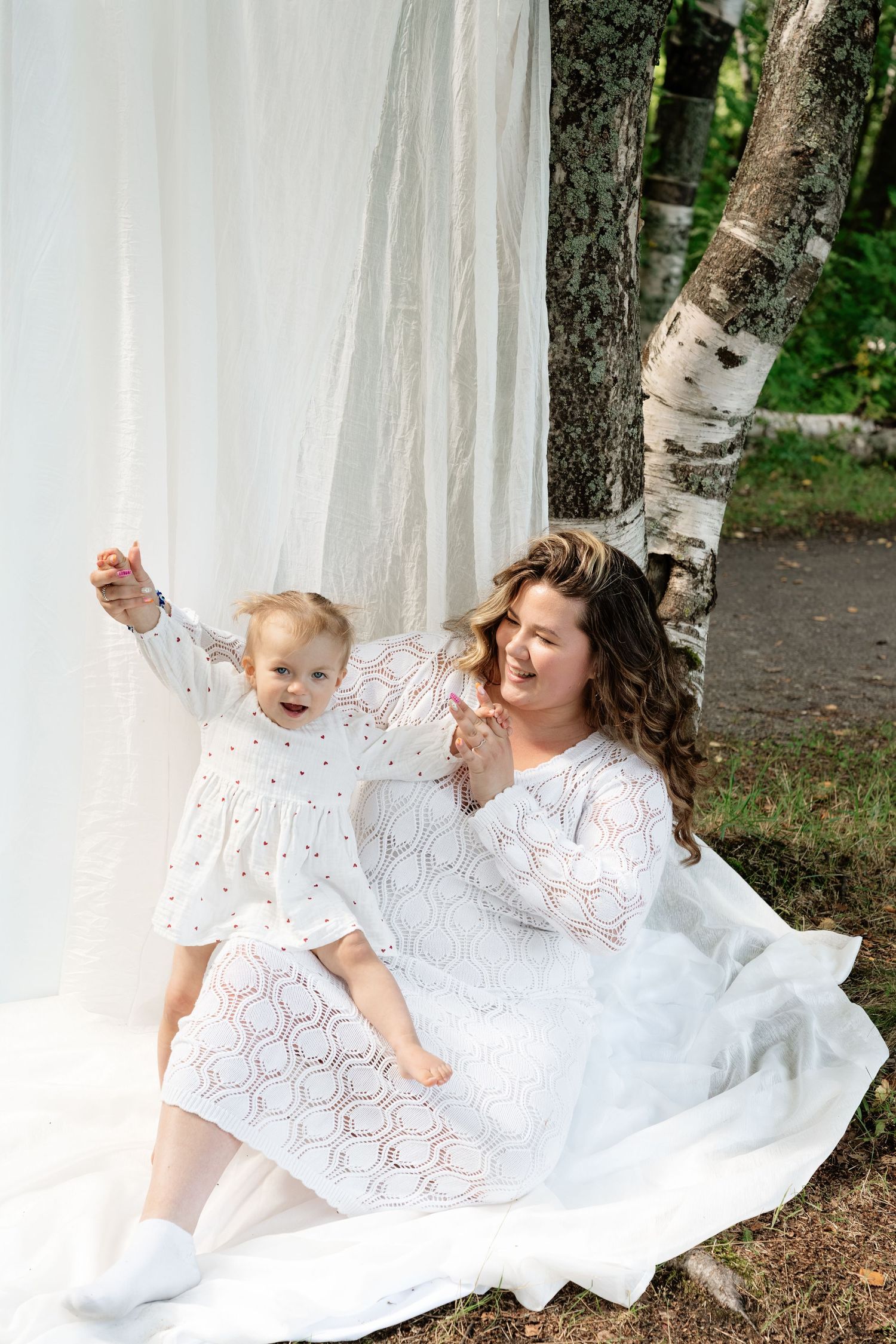 Mother and toddler in matching white dresses sitting together outdoors near birch trees with white fabric backdrop.