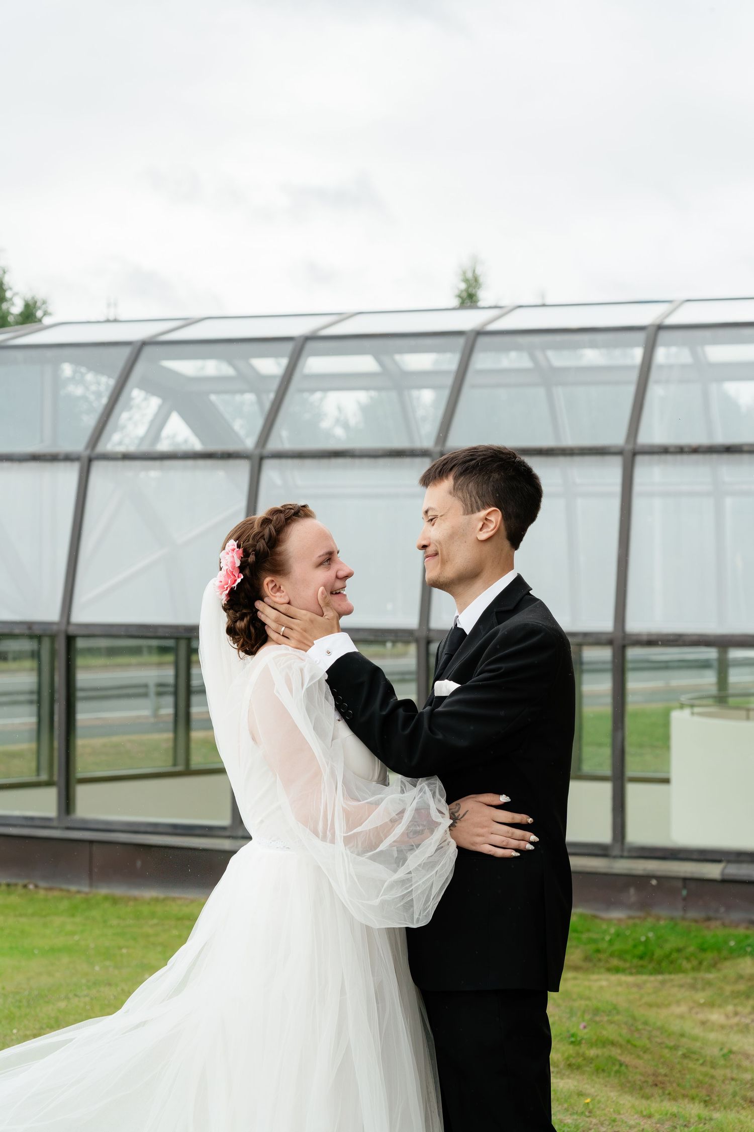 A joyful bride and groom embrace outdoors in wedding attire, smiling at each other with a glass greenhouse in the background.