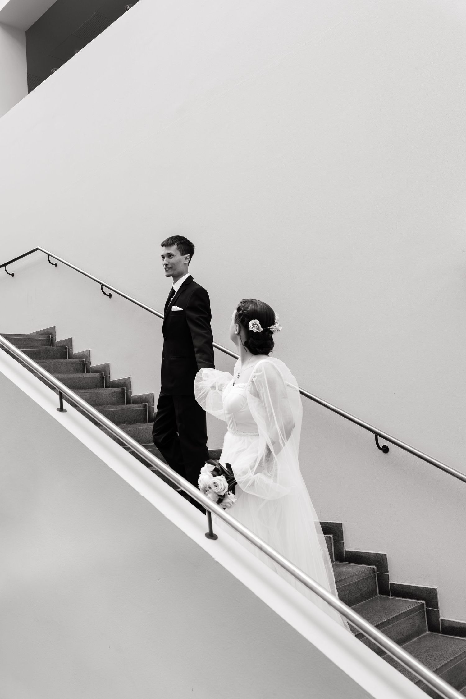 Black and white photo of bride and groom walking up stairs, groom smiling back at bride holding bouquet.
