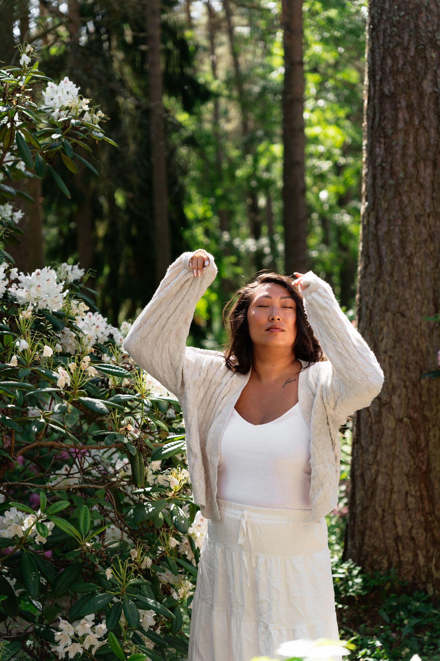 Woman in white outfit poses near blooming white rhododendron bush in sunlit forest setting.