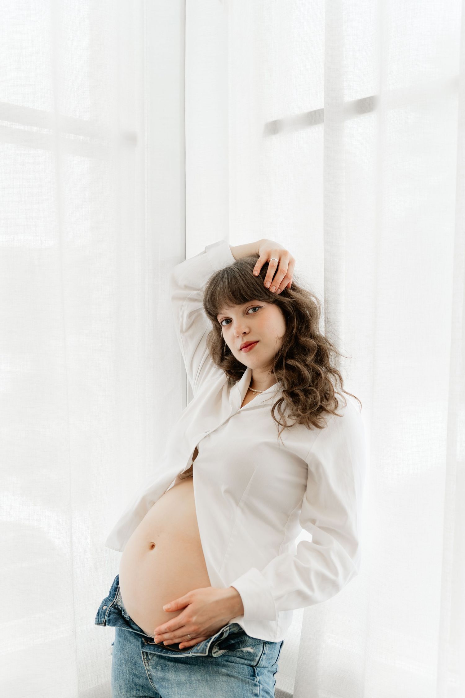 Pregnant woman in white shirt and jeans leaning against white curtains touching her belly.