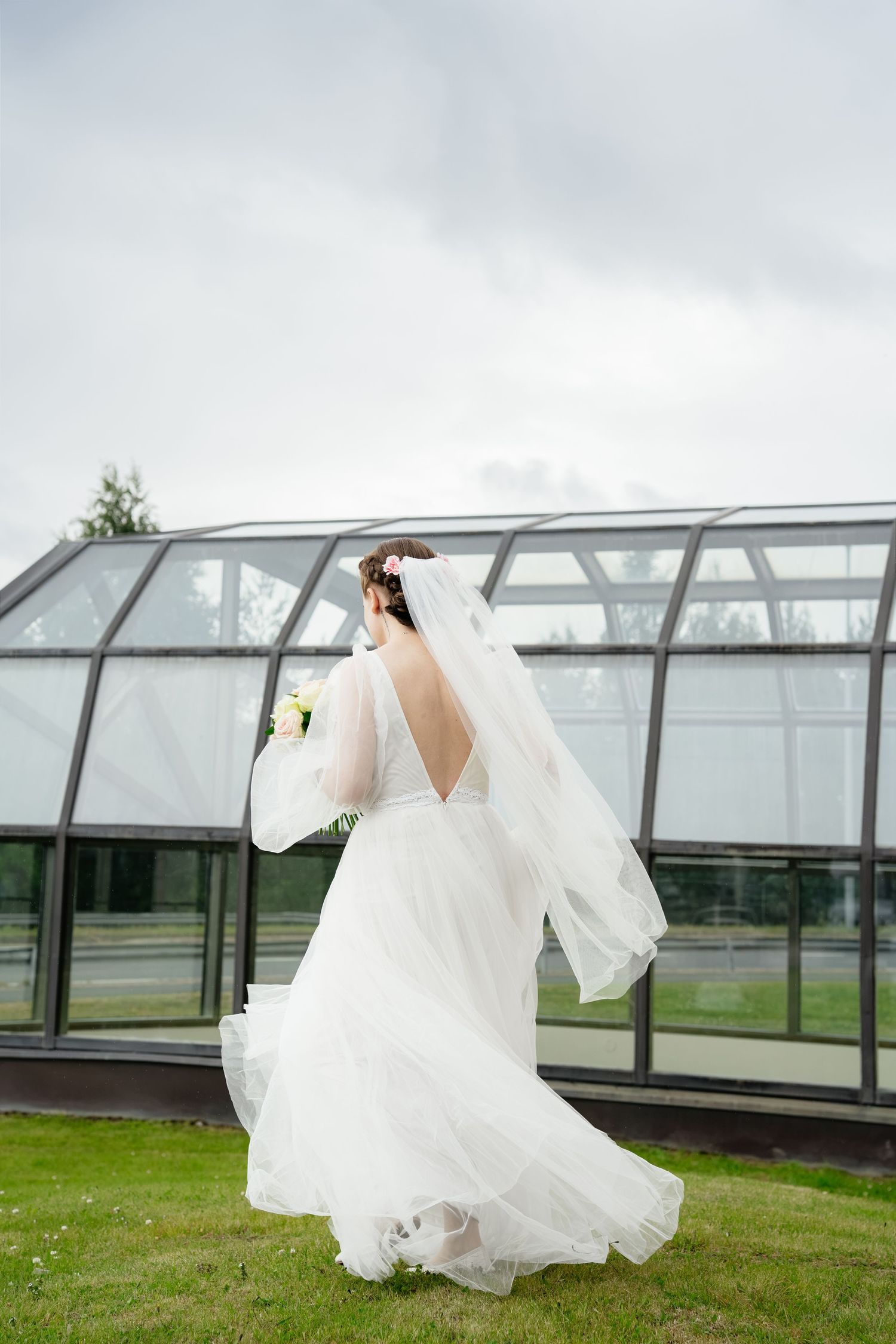Bride in white gown and veil holding bouquet, standing on green grass with glass greenhouse backdrop.
