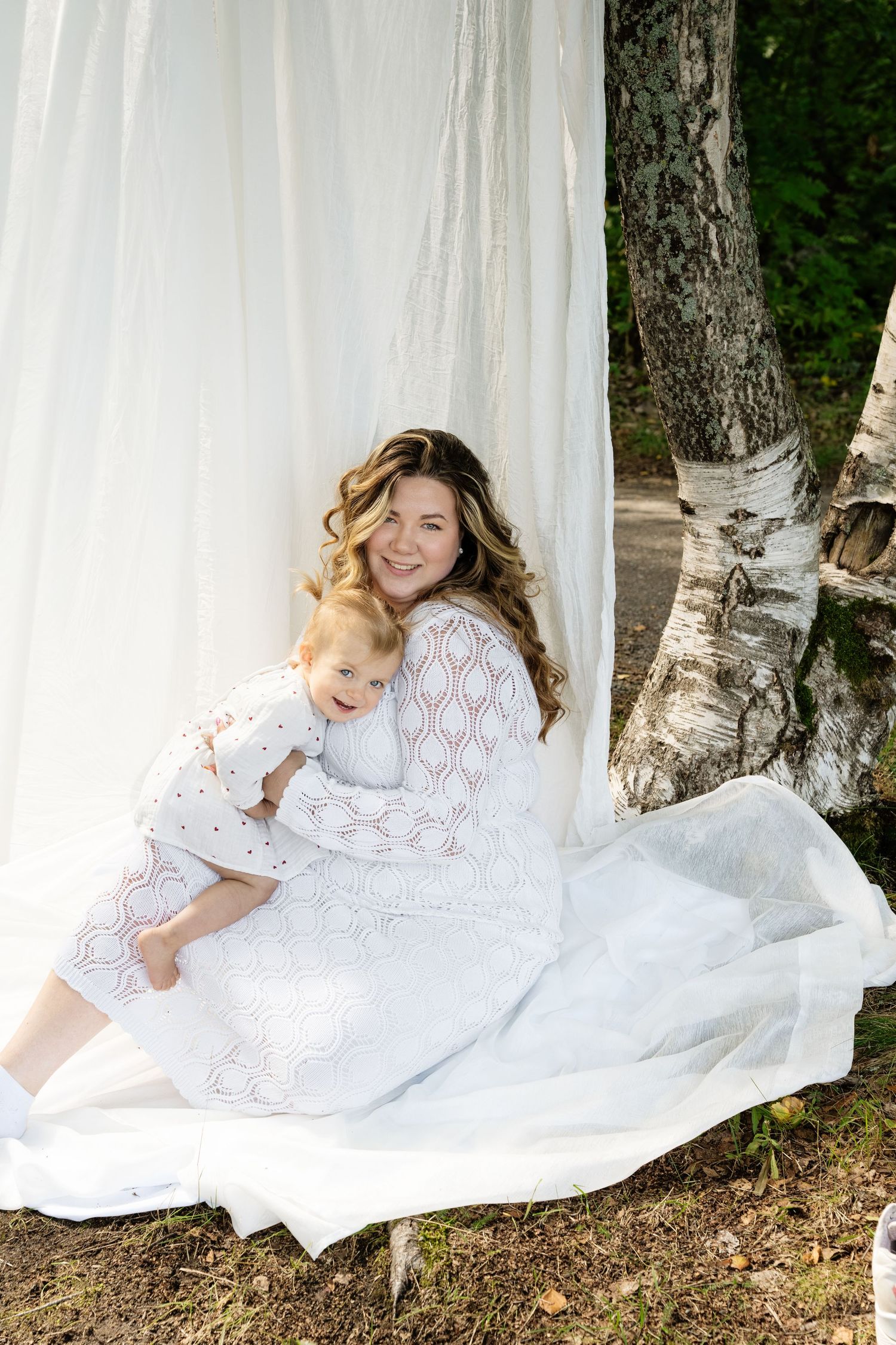 Mother in white lace dress holds toddler in white outfit outdoors near birch trees with flowing white curtain backdrop.