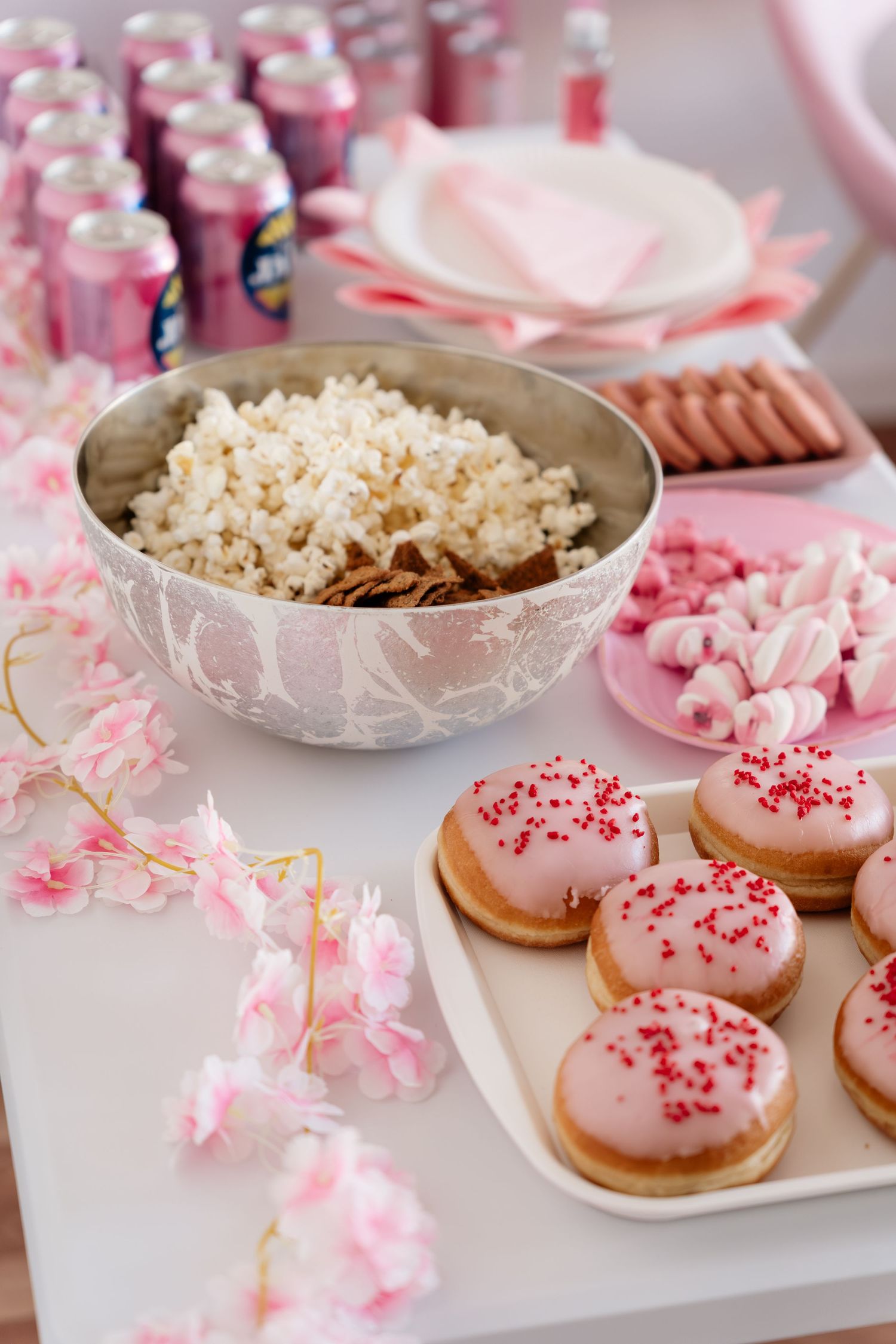 Pink party dessert table with glazed donuts, popcorn, marshmallows, and pink canned drinks.