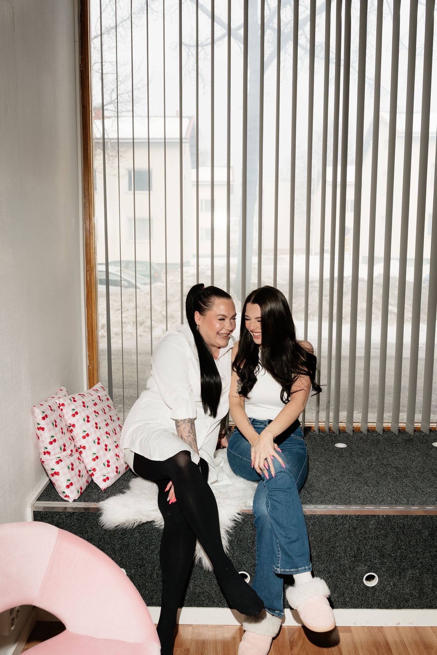 Two women with dark hair laughing together on a window bench in a bright modern interior.