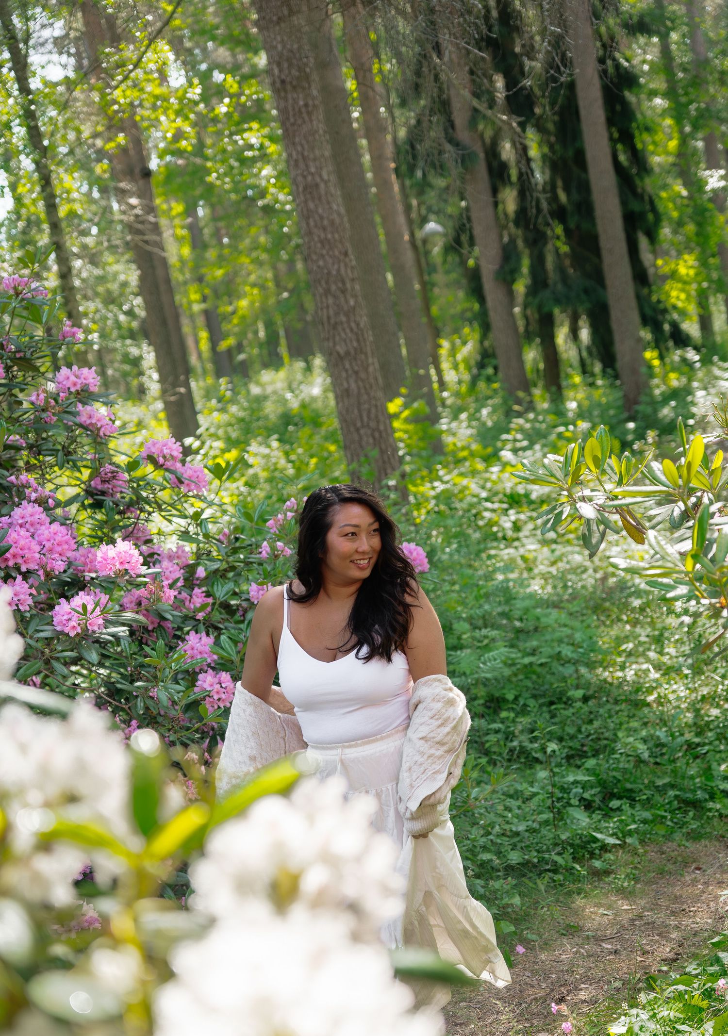 Woman in white top walks among pink rhododendrons and lush green trees on a sunny forest path.