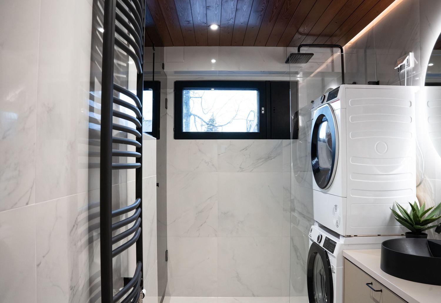 Modern laundry room with stacked washer-dryer, marble walls, black heated towel rail, wooden ceiling, and small window.