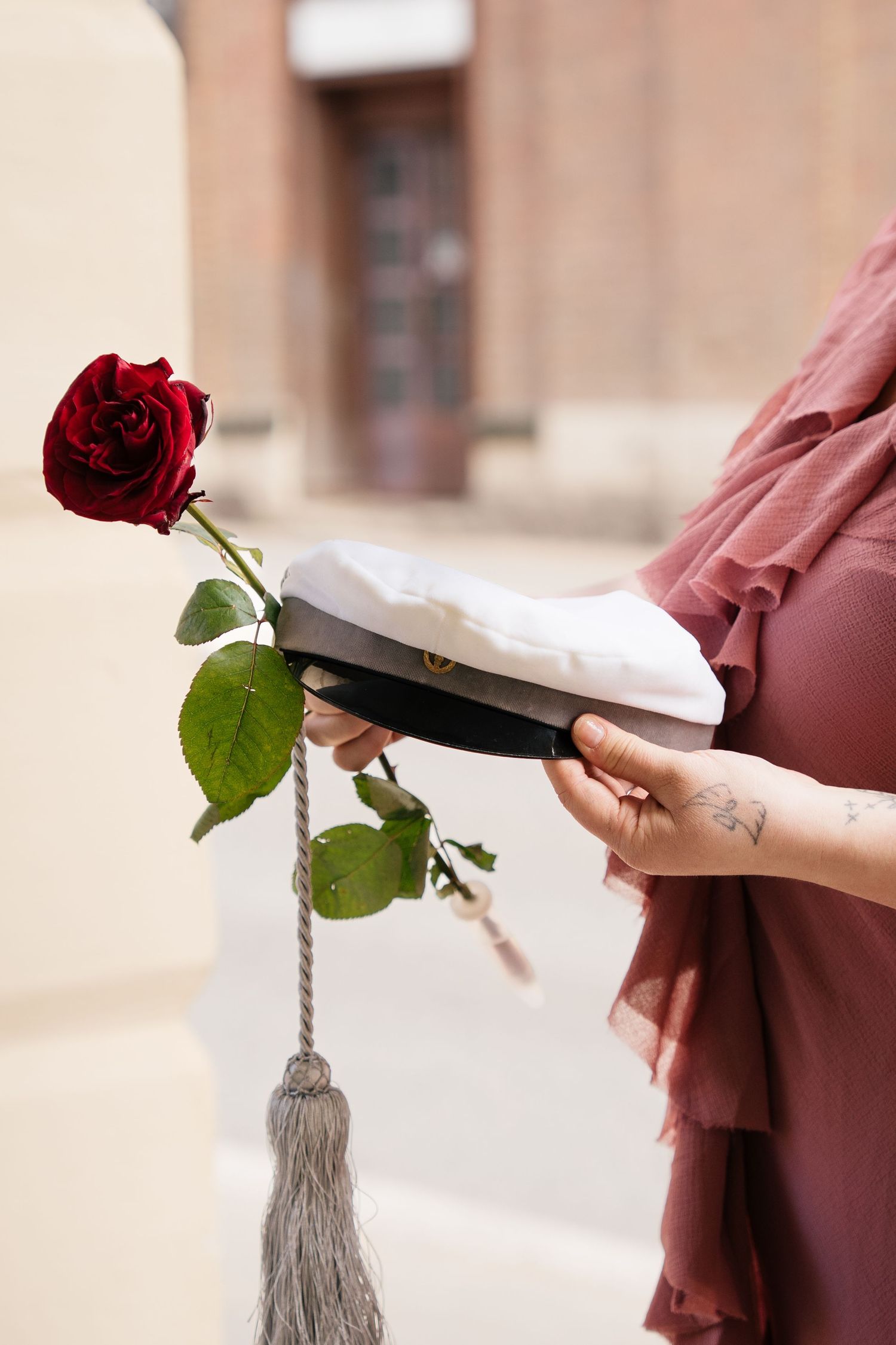 Woman in pink dress holding graduation cap with red rose and tassel outdoors.